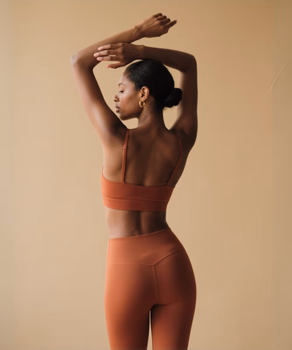 Woman in rust-colored activewear with arms raised, posing sideways against a beige background.
