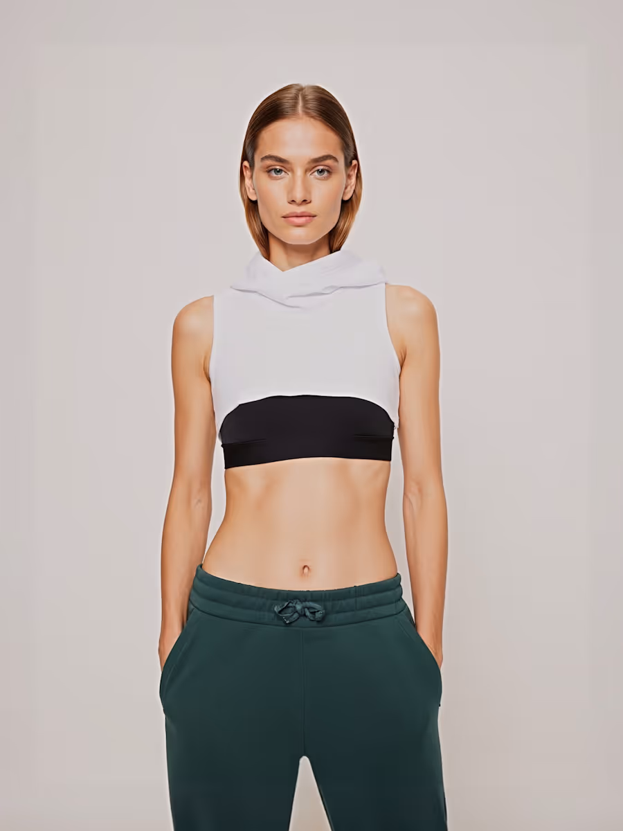 Woman wearing a white cropped sleeveless hoodie over a black sports bra and dark green sweatpants with hands in pockets.