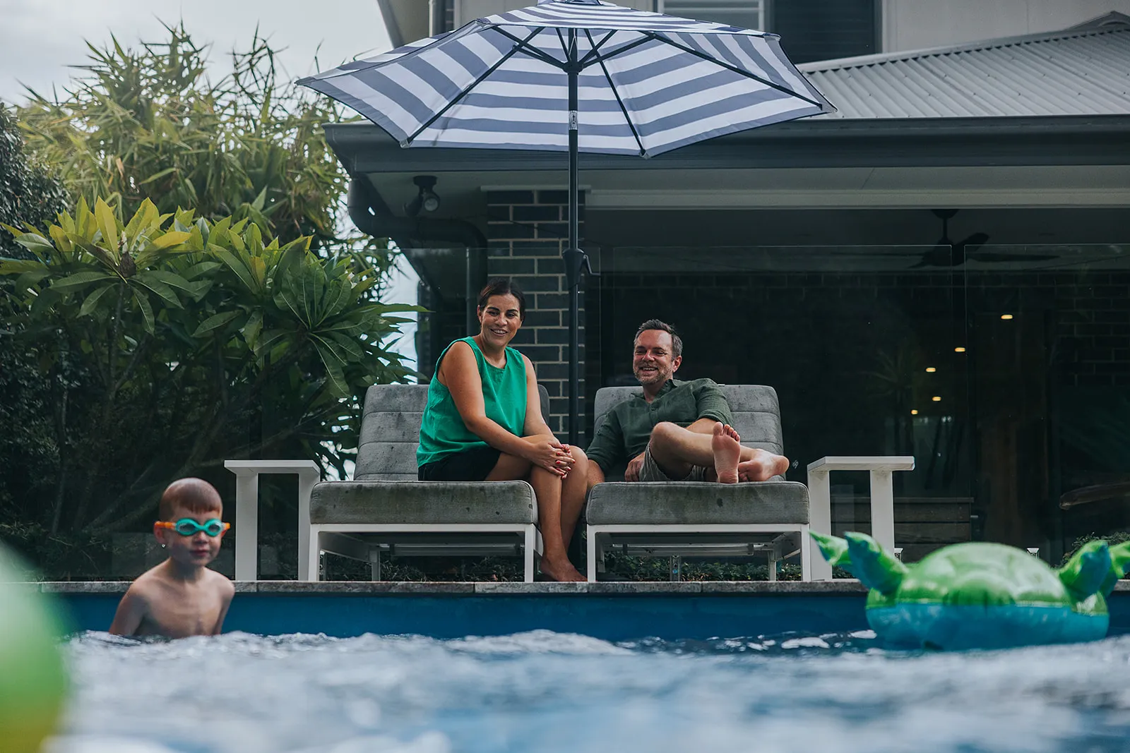 Couple relaxing on poolside lounge chairs under umbrella while child swims nearby wearing goggles.
