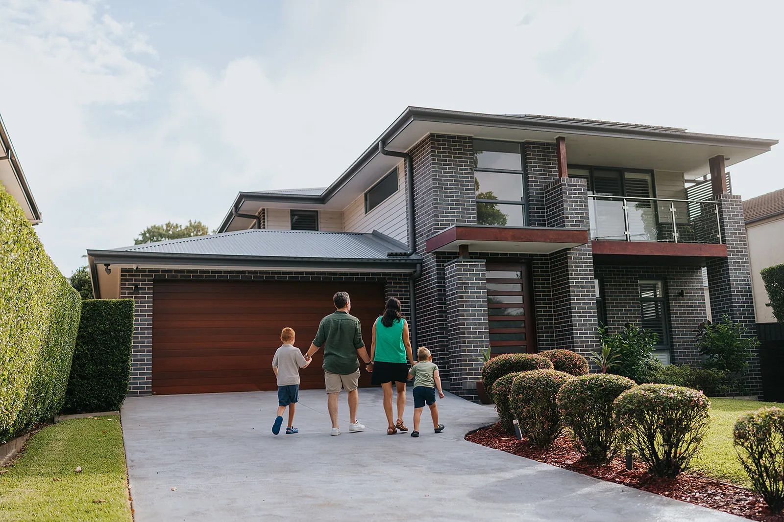 Family of four walking hand in hand toward a modern two-story brick house with a wooden garage door.