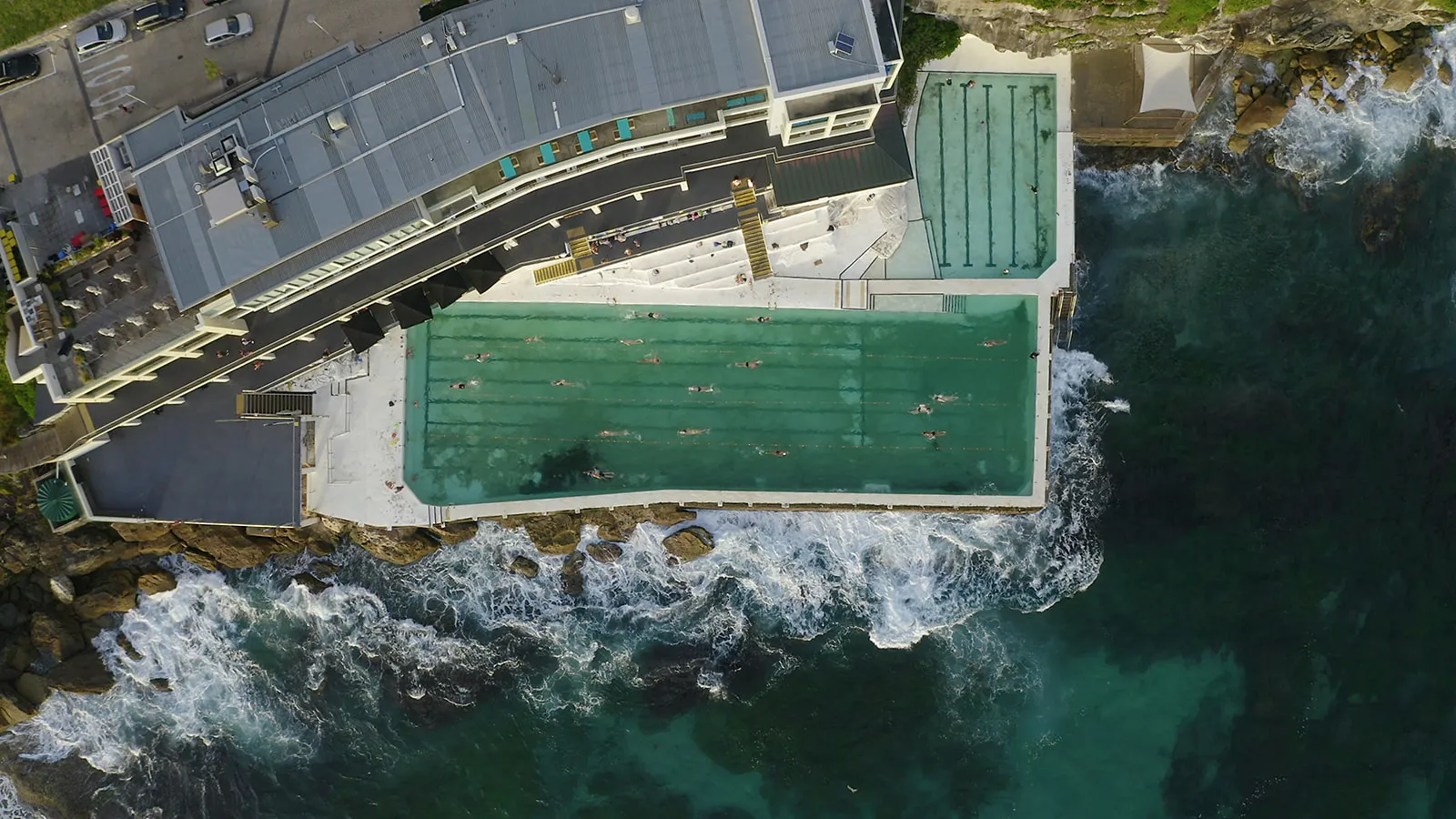Aerial view of a curved ocean pool next to waves crashing on rocks with swimmers inside.