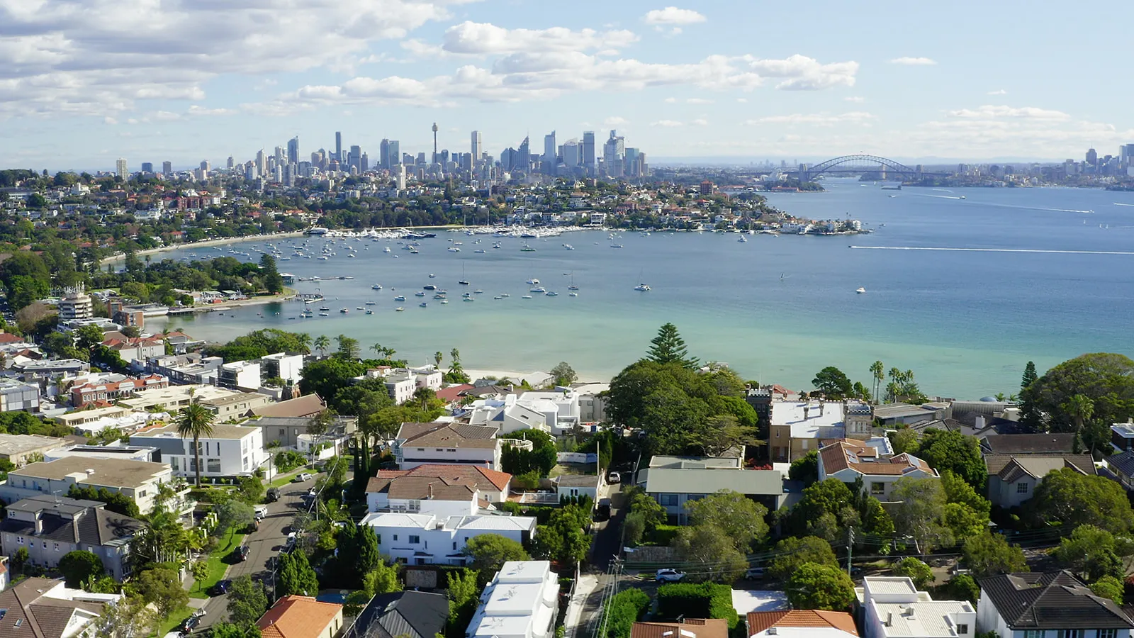 Aerial view of Sydney Harbour with boats, residential area, and city skyline with Harbour Bridge.
