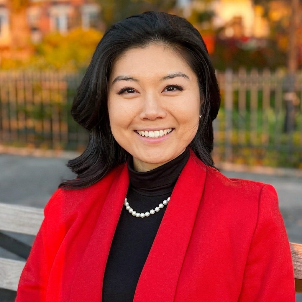 Odelia Xing,smiling. Woman with black hair wearing a red blazer, black turtleneck, and pearl necklace sitting on a bench outdoors.