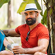 Smiling man with beard wearing a light blue fedora and red shirt holding a phone indoors with green plants in the background.