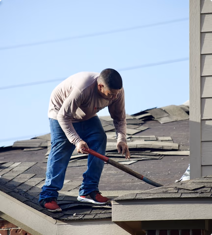 Man wearing red and black shoes, jeans, and beige long-sleeve shirt working on a roof with a crowbar to remove old shingles.