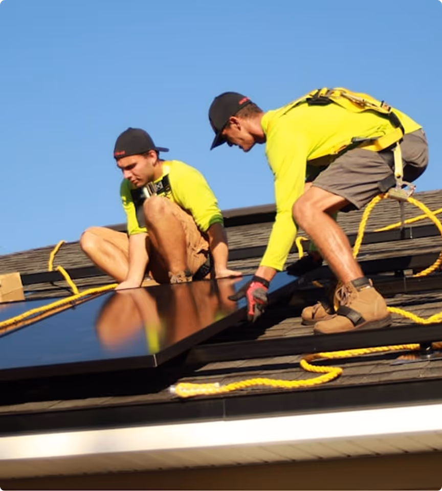 Two workers wearing yellow shirts and safety harnesses installing solar panels on a rooftop under clear blue sky.