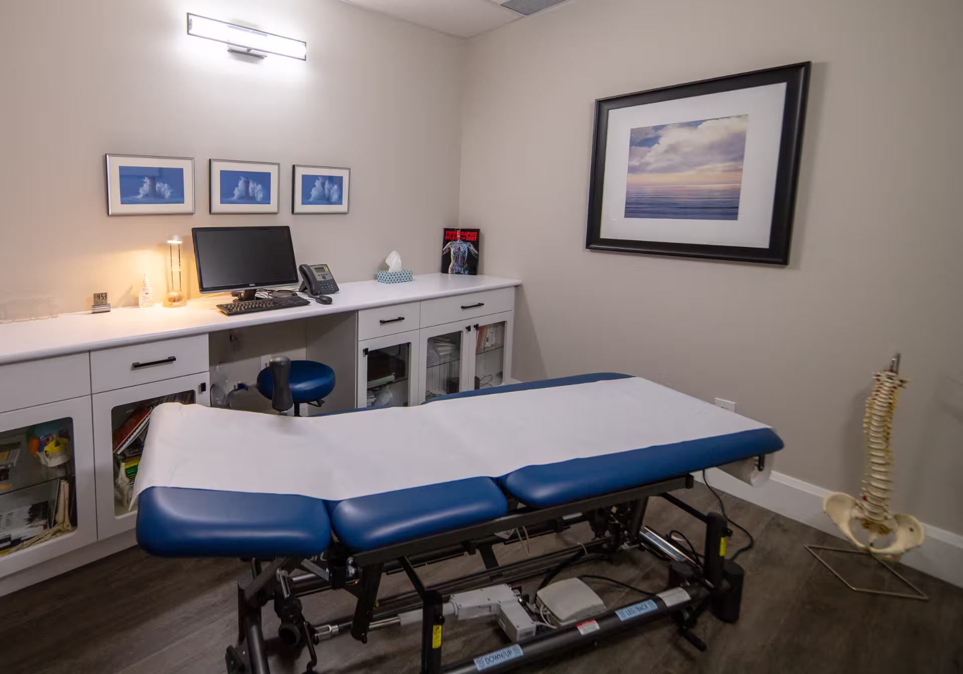 Medical examination room with a blue and white treatment table, a desk with computer and phone, and a spine model on the floor.