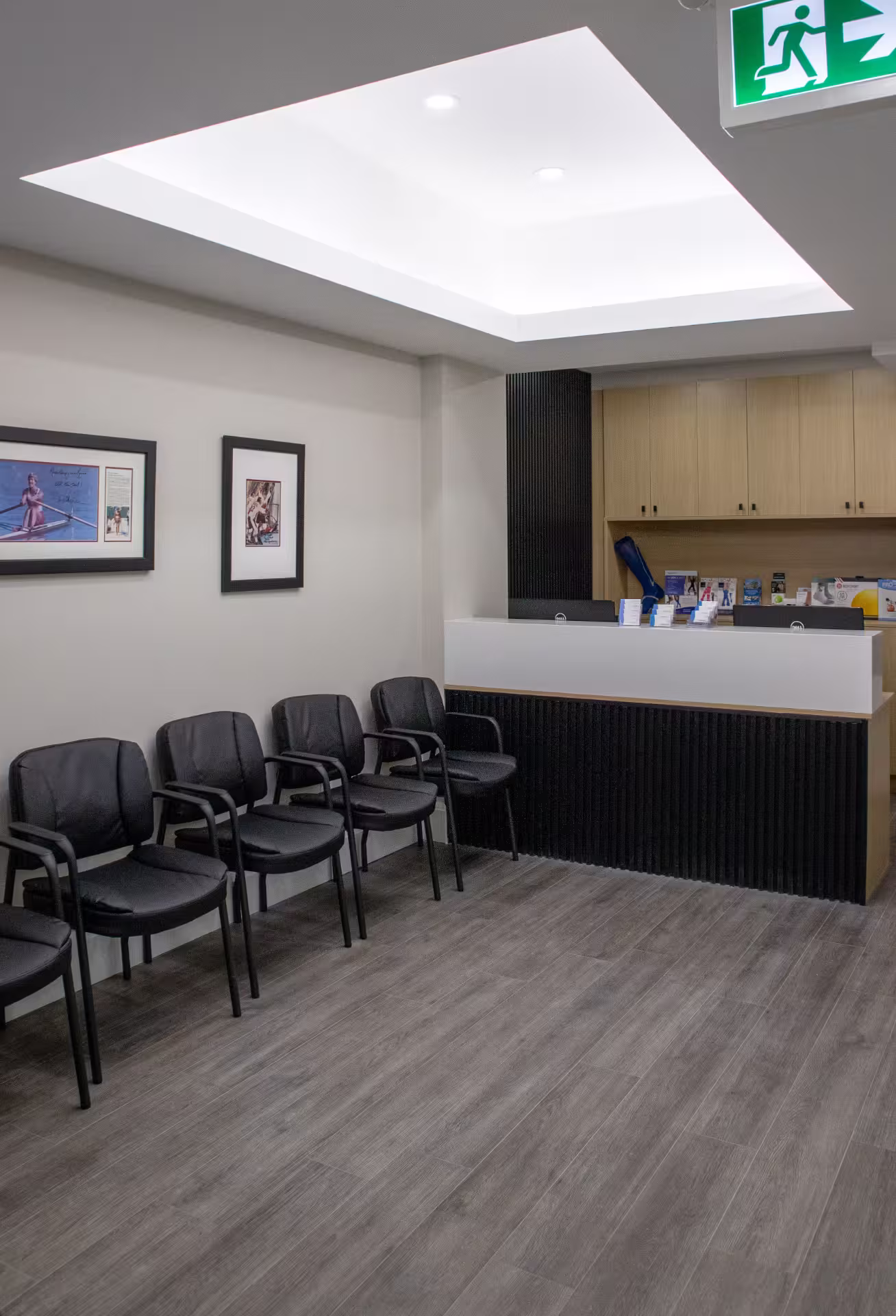 Modern waiting room with black chairs aligned against a wall, a reception desk, framed pictures, and an illuminated exit sign.