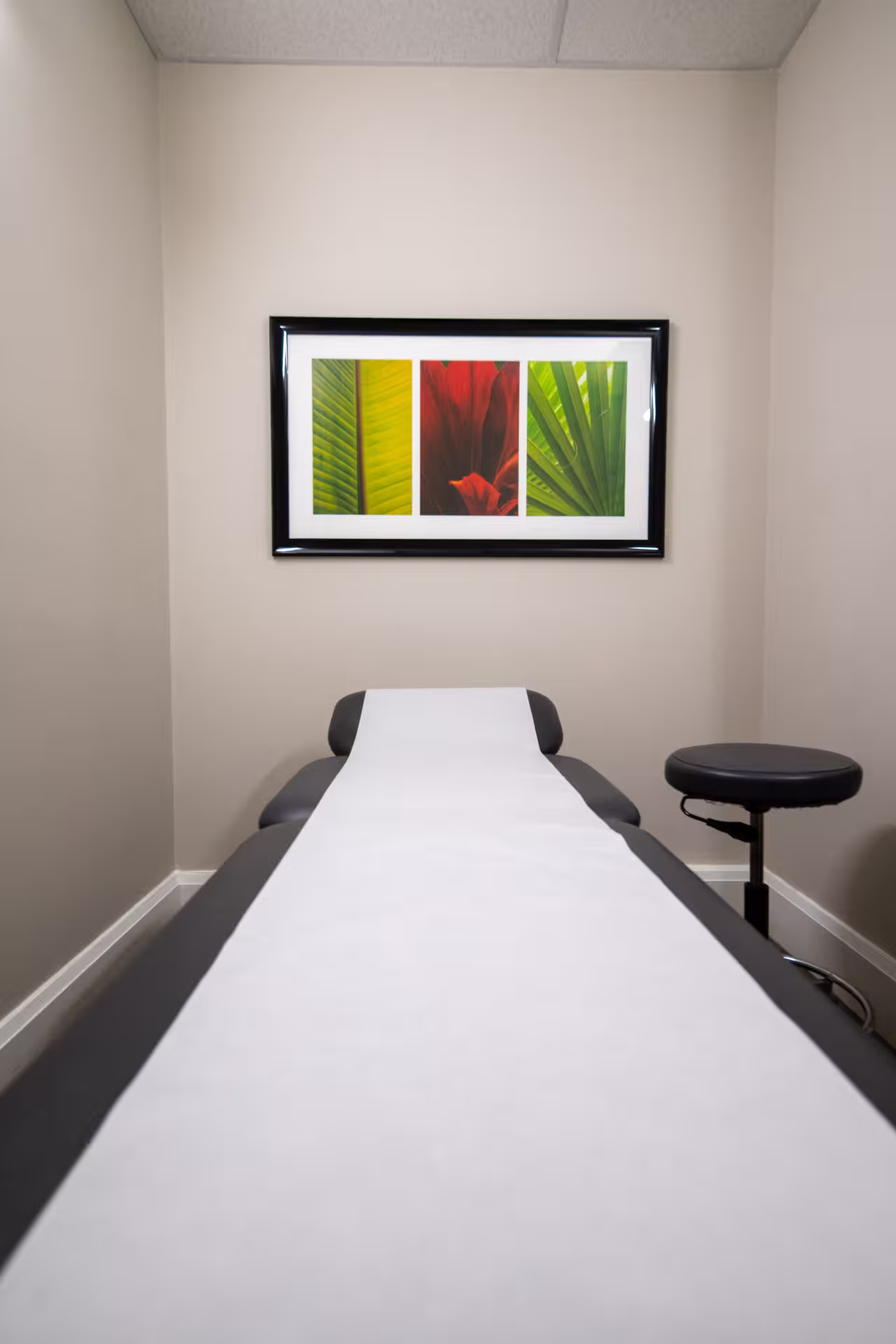 Examination room with a black medical table covered in white paper and a black stool beside it, featuring a framed artwork of three colorful leaf sections on the beige wall.