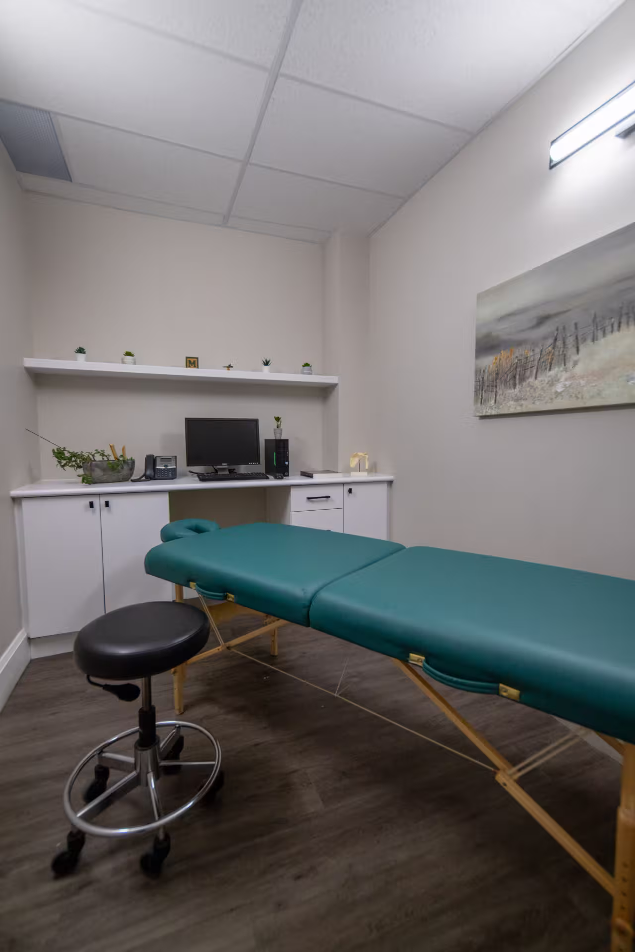 Medical examination room with a teal patient table, black rolling stool, and a desk with a computer and small plants.