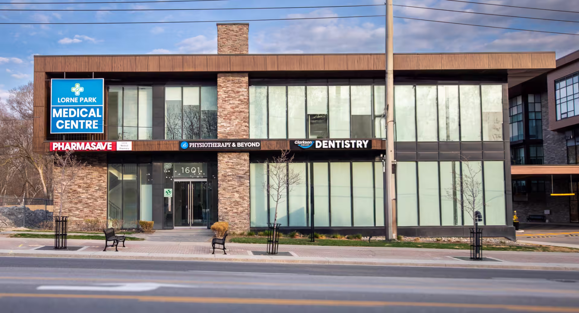 Modern two-story building housing Lorne Park Medical Centre, Pharmasave, Physiotherapy & Beyond, and Clarkson Dental with large windows and benches outside.