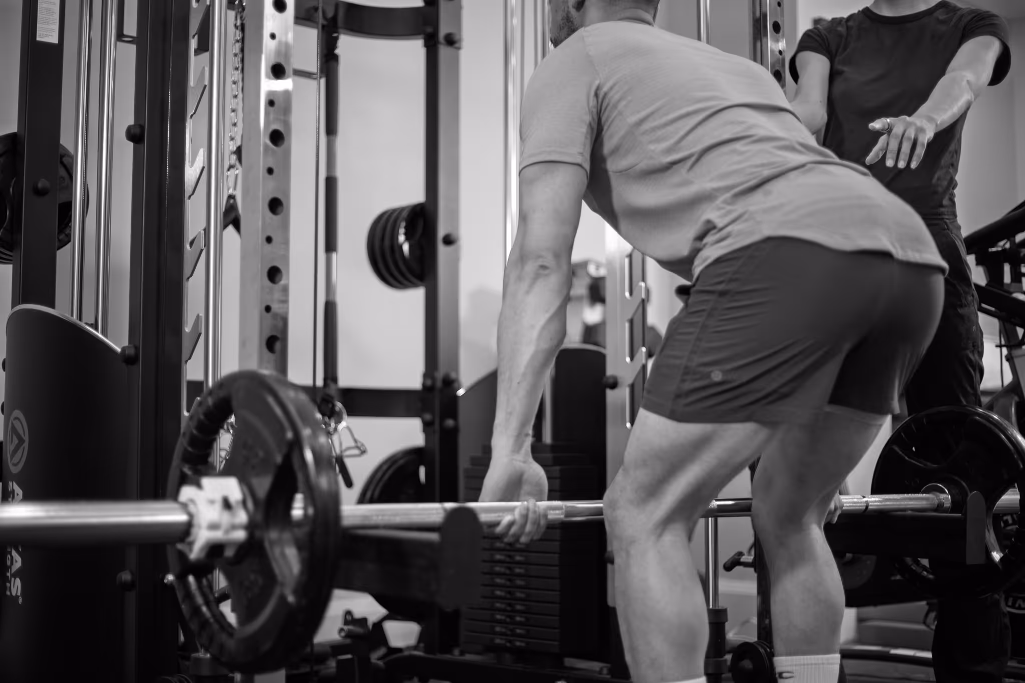 Man lifting a barbell with weights in a gym while a trainer guides him.