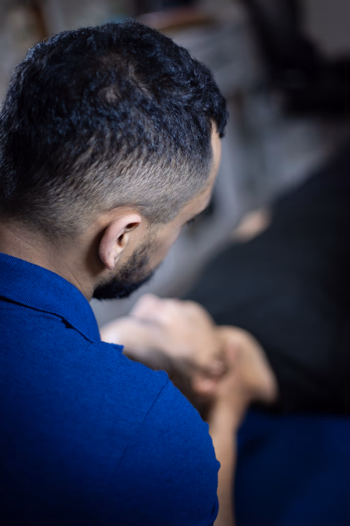 Man with short hair in blue shirt looking down with hands clasped, viewed from behind.