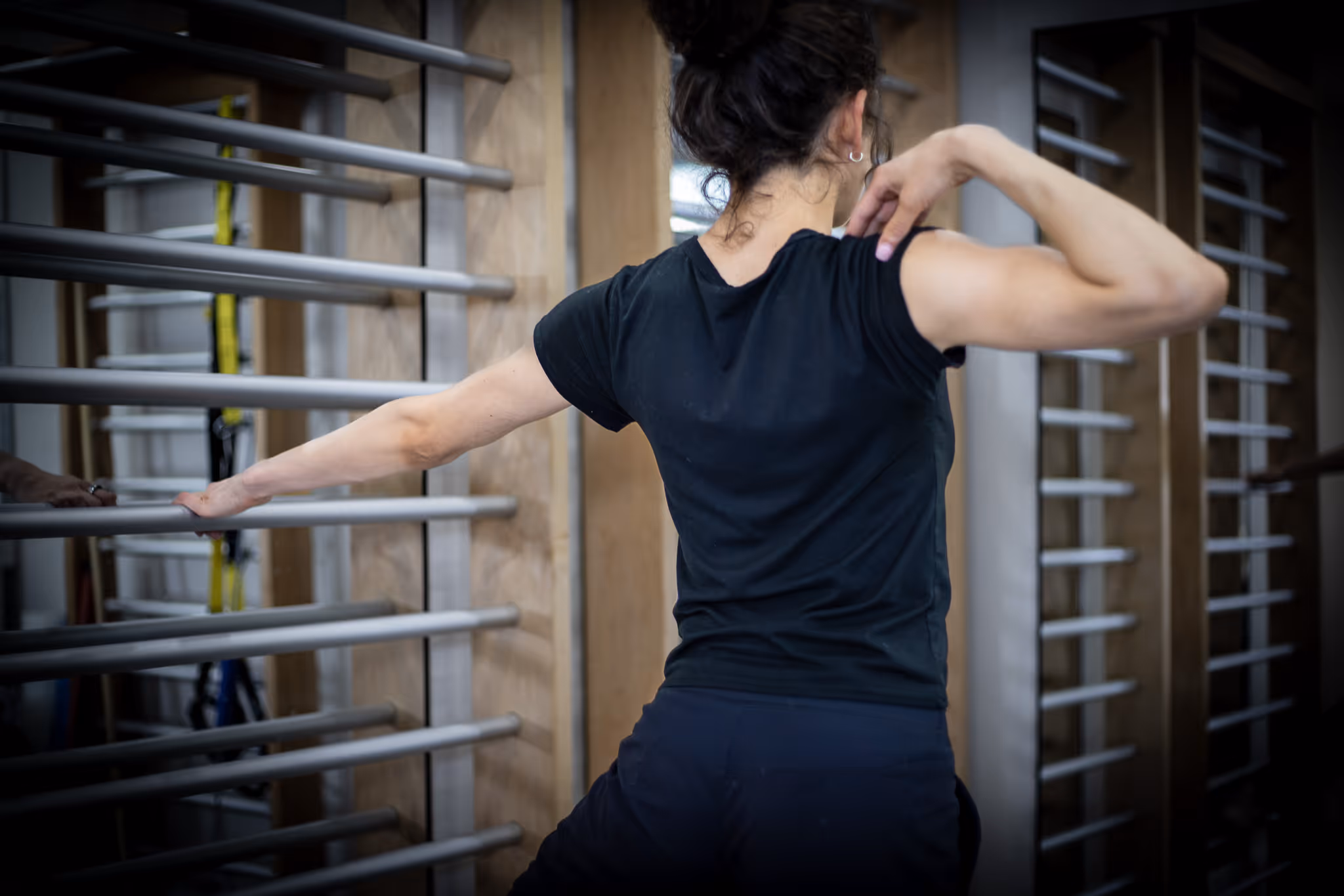 Woman stretching her arm while holding a metal bar in a gym with wooden wall bars.