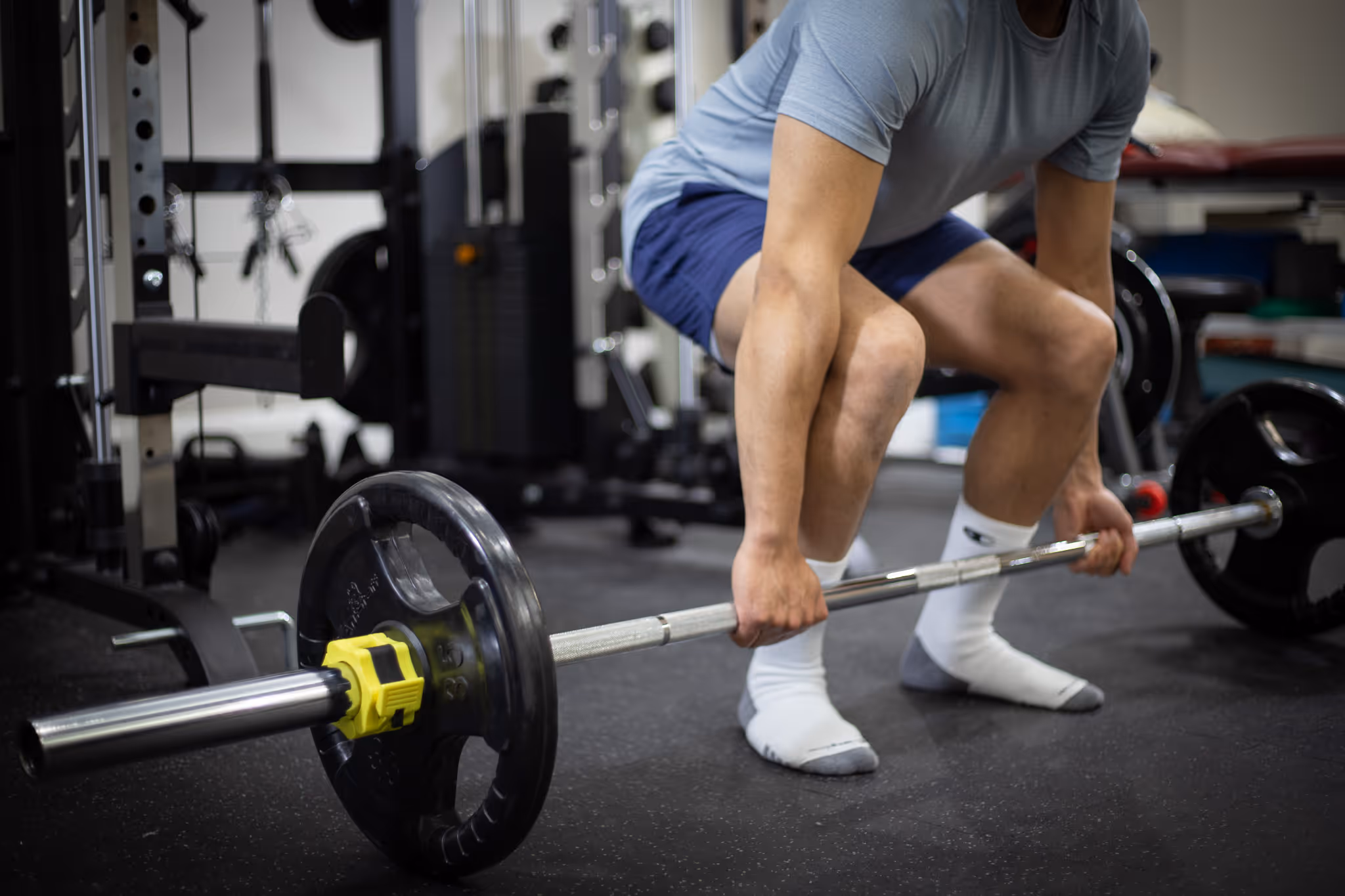Person in socks lifting a barbell with black weight plates in a gym setting.