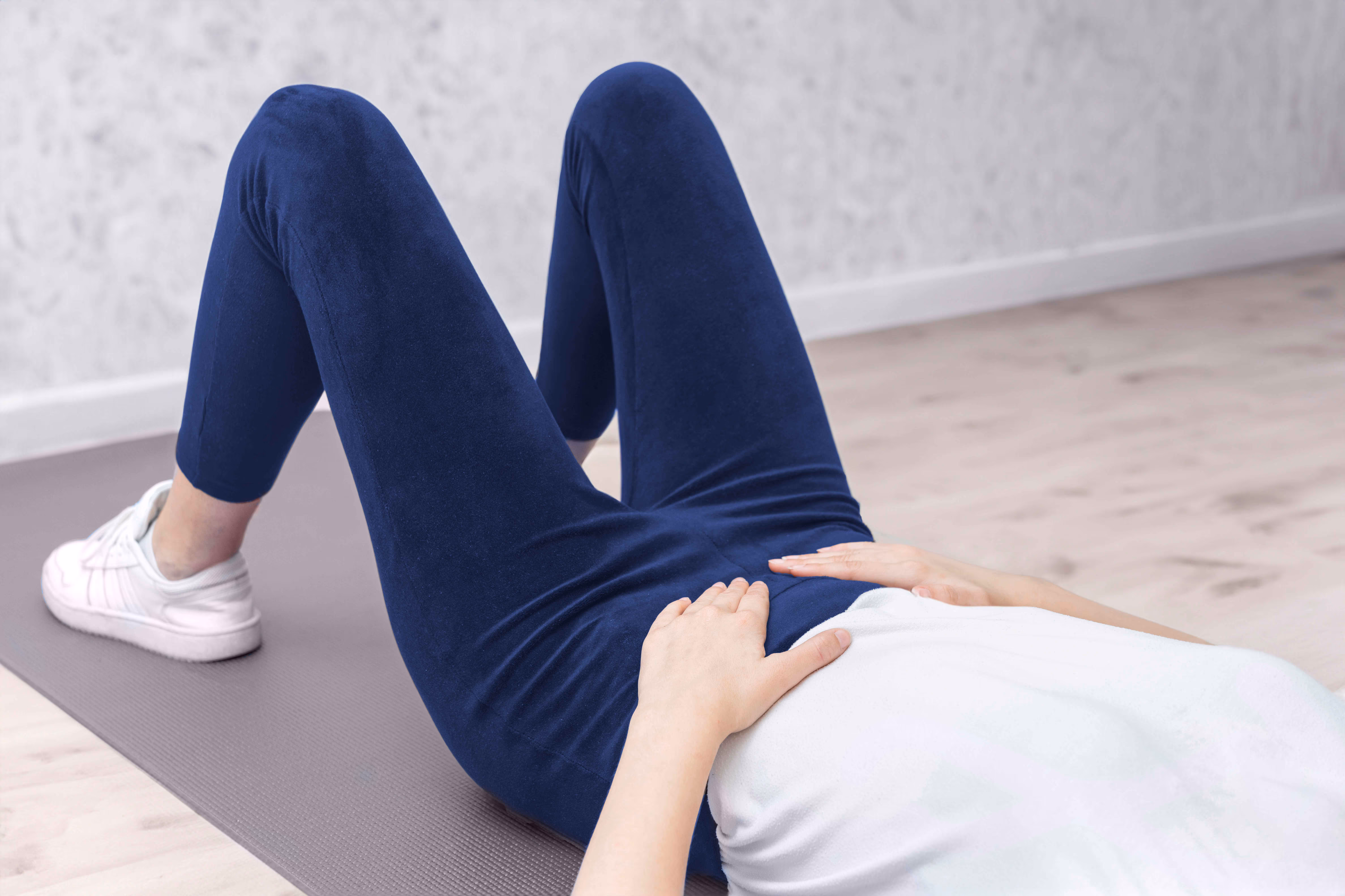 Person lying on exercise mat with knees bent, hands resting on stomach, wearing navy leggings and white sneakers.