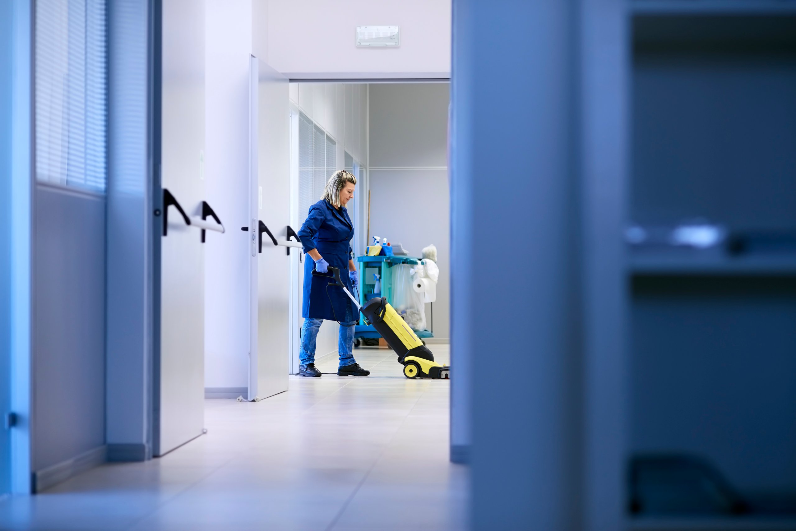 Janitor cleaning hallway floor with a yellow vacuum cleaner wearing a blue uniform and gloves.