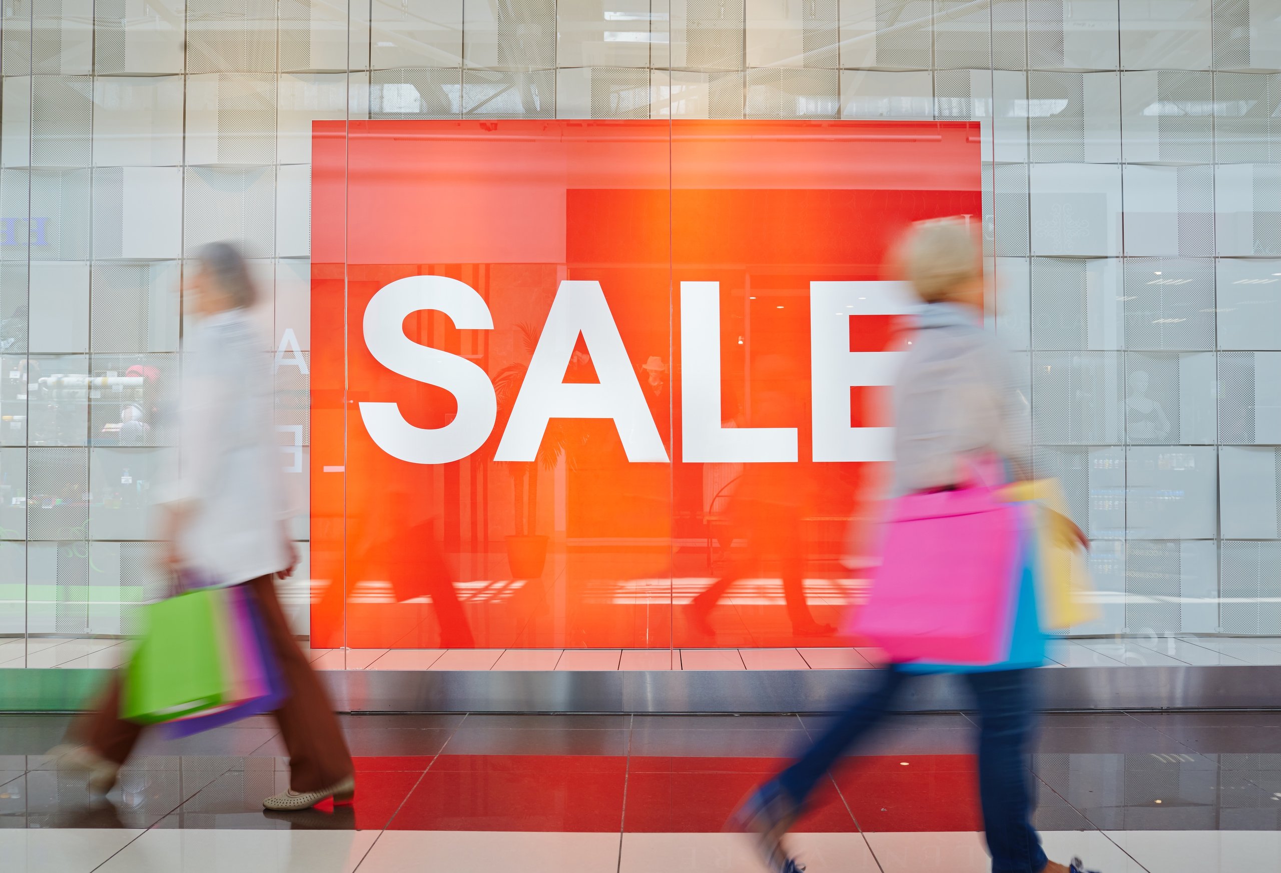 Two blurred shoppers carrying colorful bags walking past a large red sale sign in a mall.
