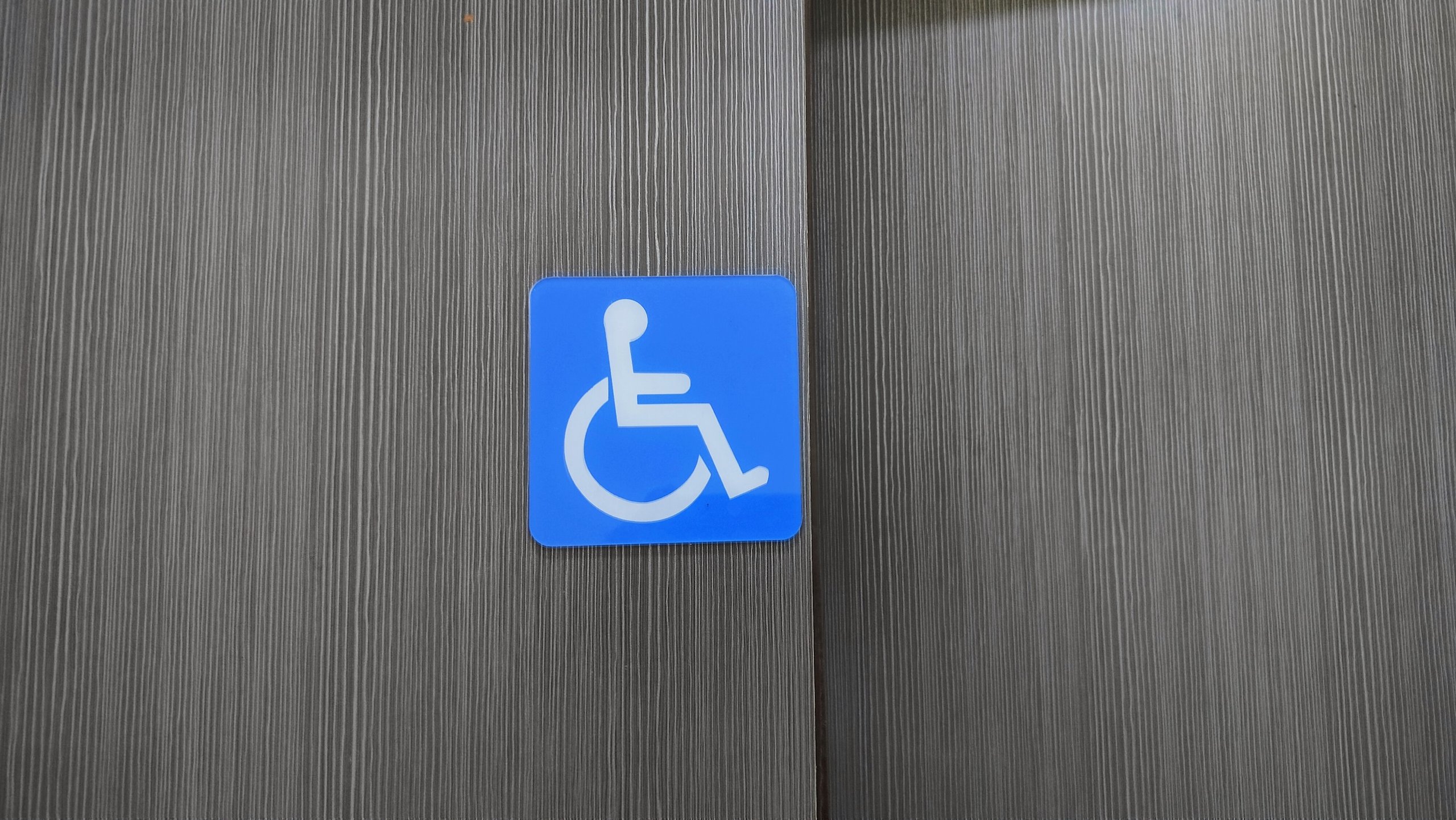 Blue accessibility sign with white wheelchair symbol mounted on a vertically striped gray wooden wall.