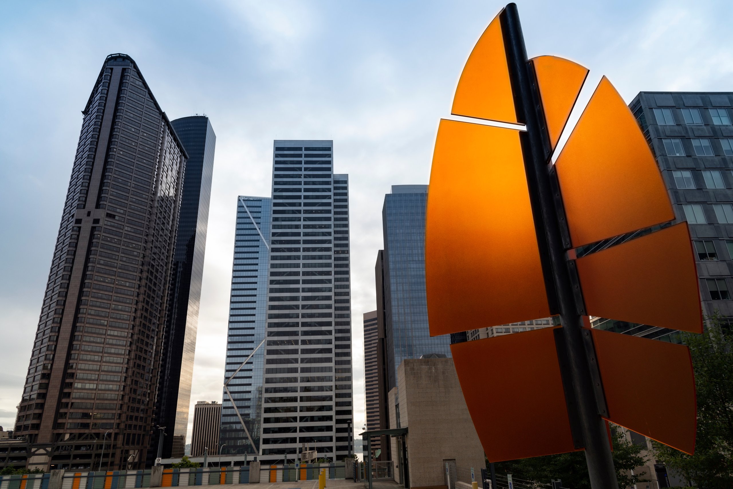Orange modern sculpture with segmented panels in front of tall glass and steel skyscrapers under a cloudy sky.