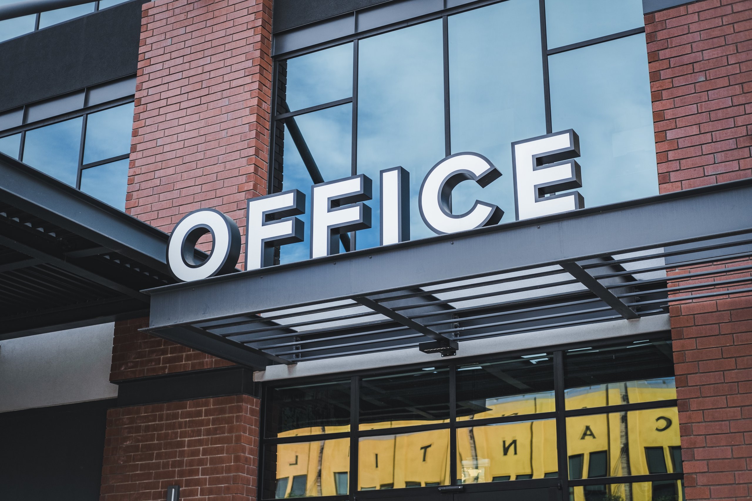 Modern office building entrance with large white letters spelling 'OFFICE' above glass doors and brick walls.