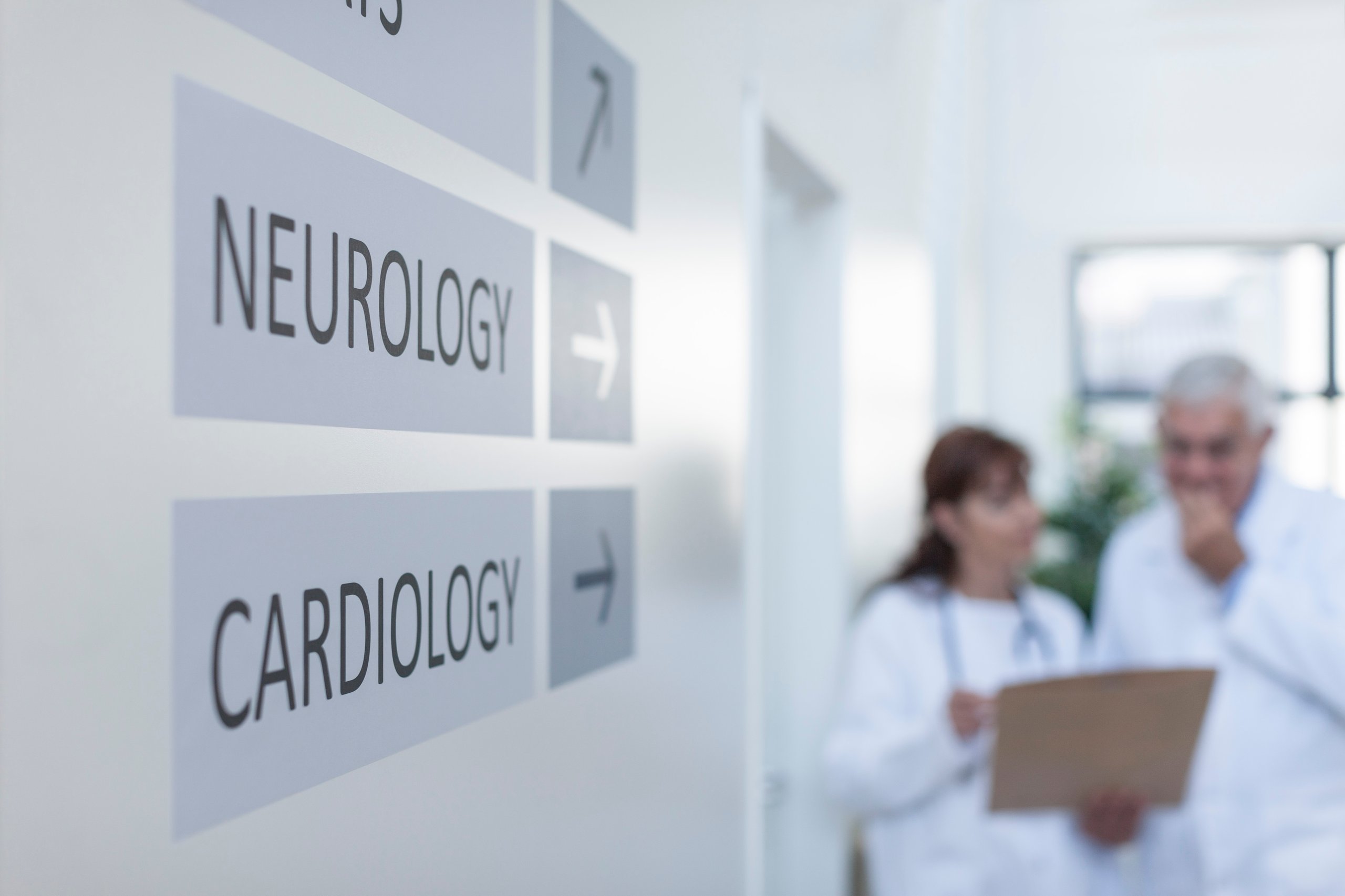 Hospital hallway sign pointing to Neurology and Cardiology departments with two doctors discussing a clipboard in the background.