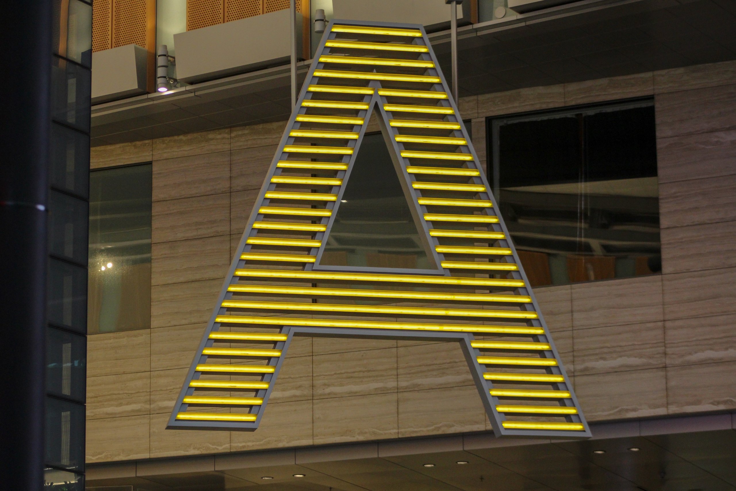 Large illuminated letter A with horizontal yellow light strips mounted on a building facade.