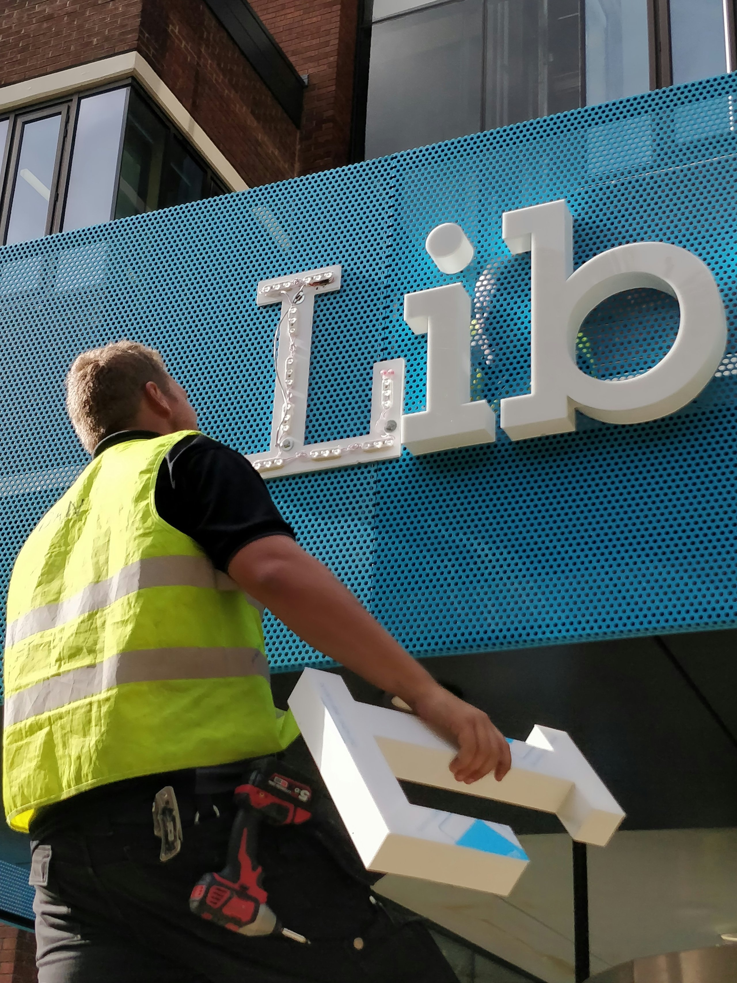 Worker in yellow safety vest holding a large white letter 'L' while installing illuminated building signage reading 'Lib'.