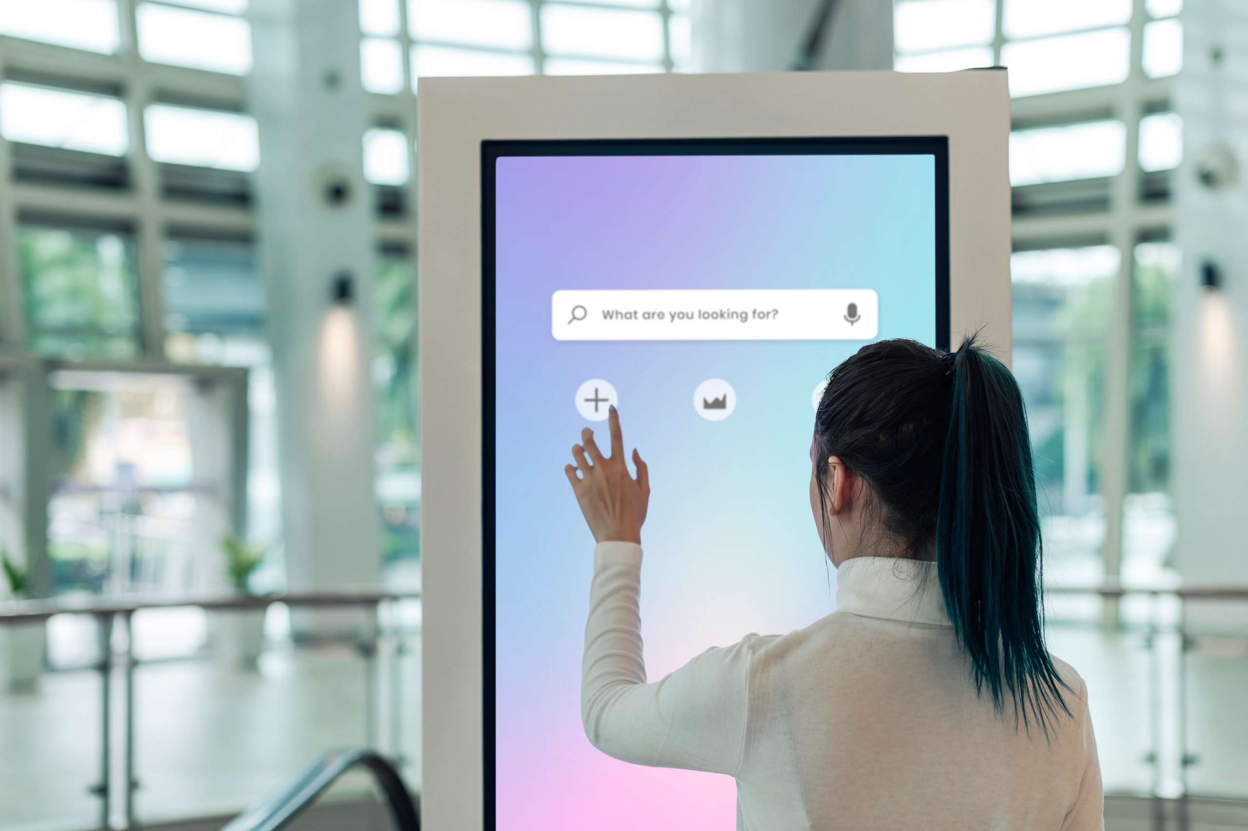 Woman with a ponytail interacting with a large touchscreen kiosk displaying a search bar and icons in a modern indoor space.