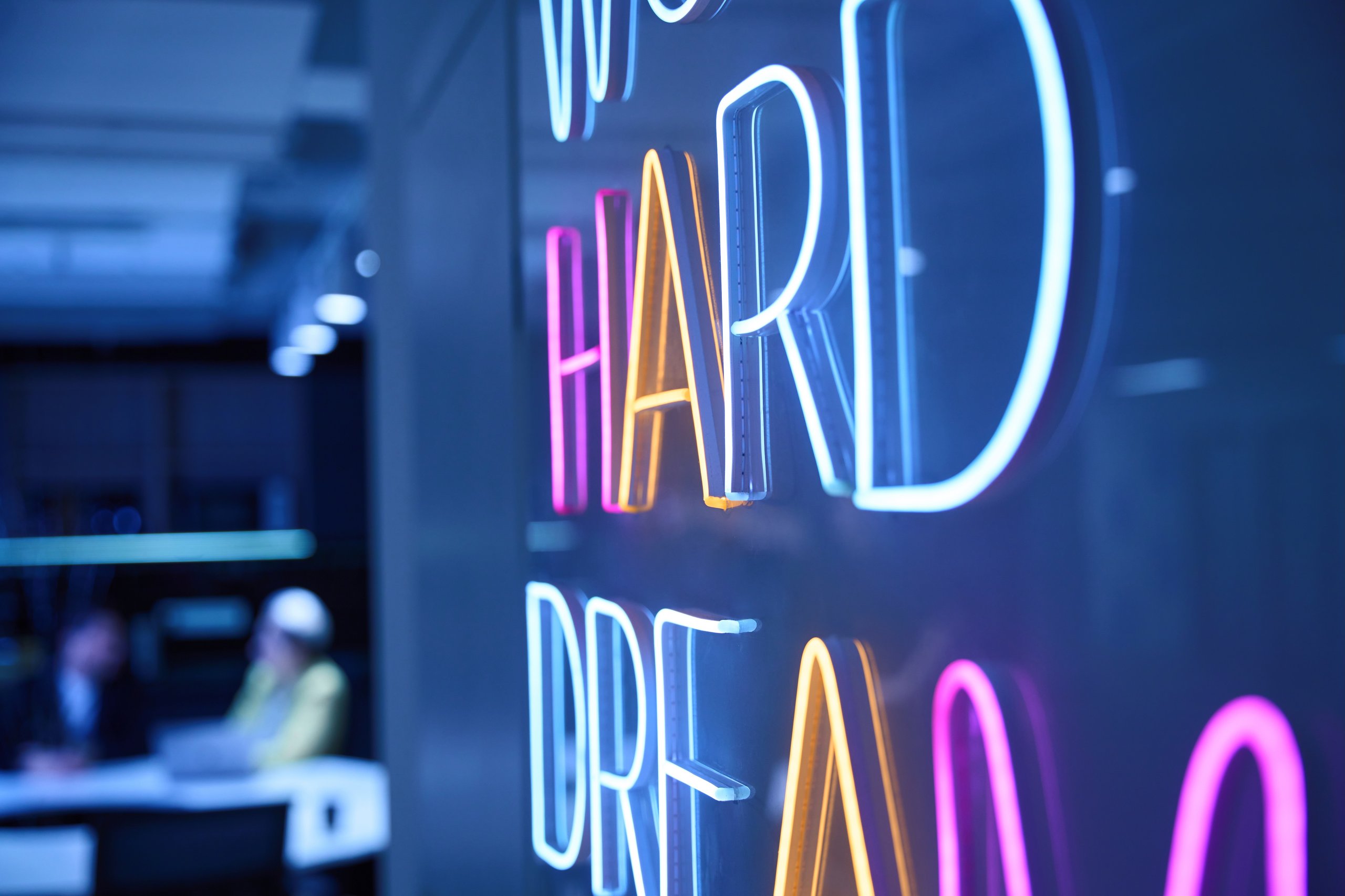 Colorful neon sign displaying the words 'HARD DREAM' on a dark wall with a blurred office background.