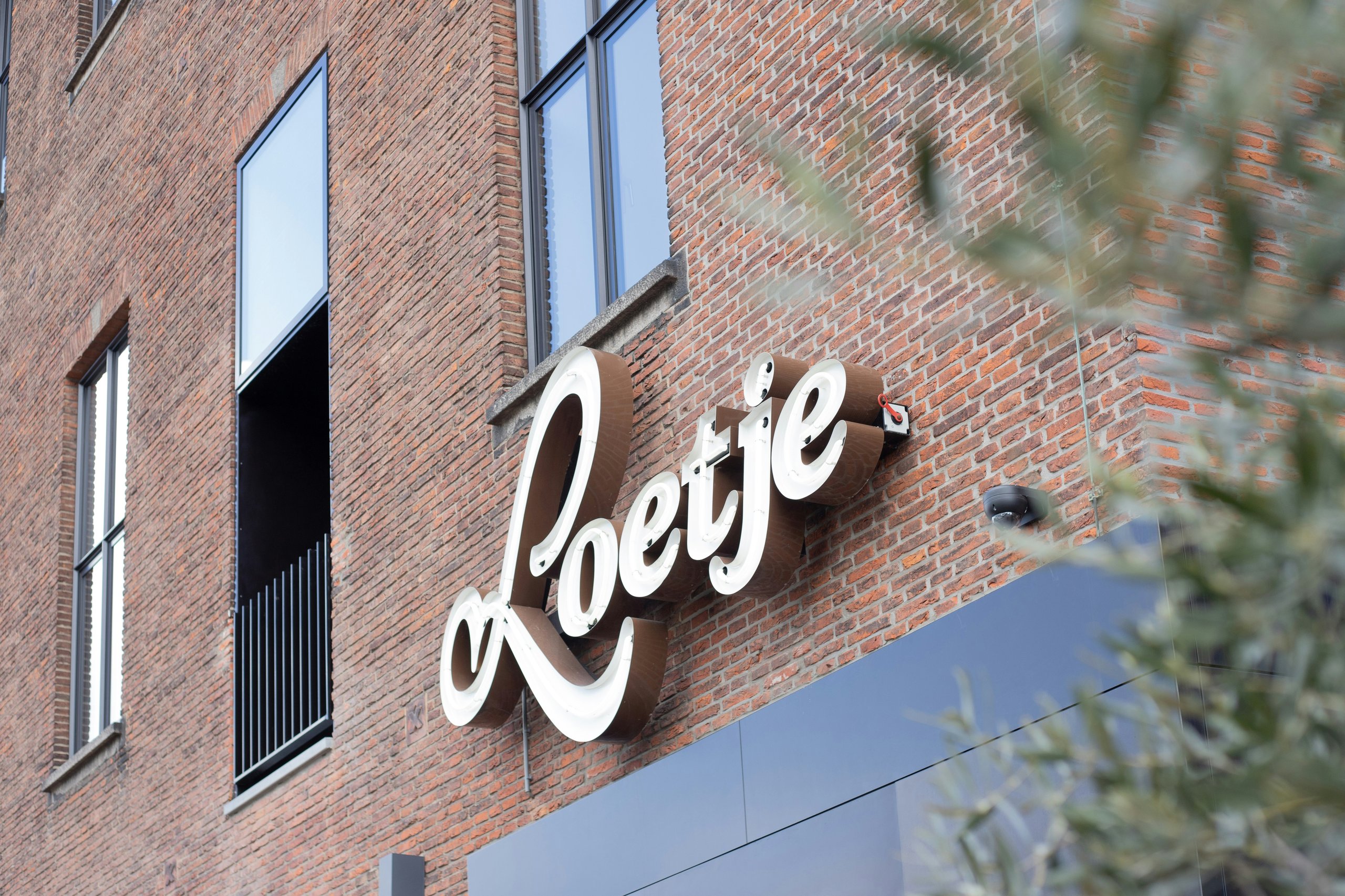 White and brown illuminated sign reading 'Loetje' mounted on a brick building exterior with windows.