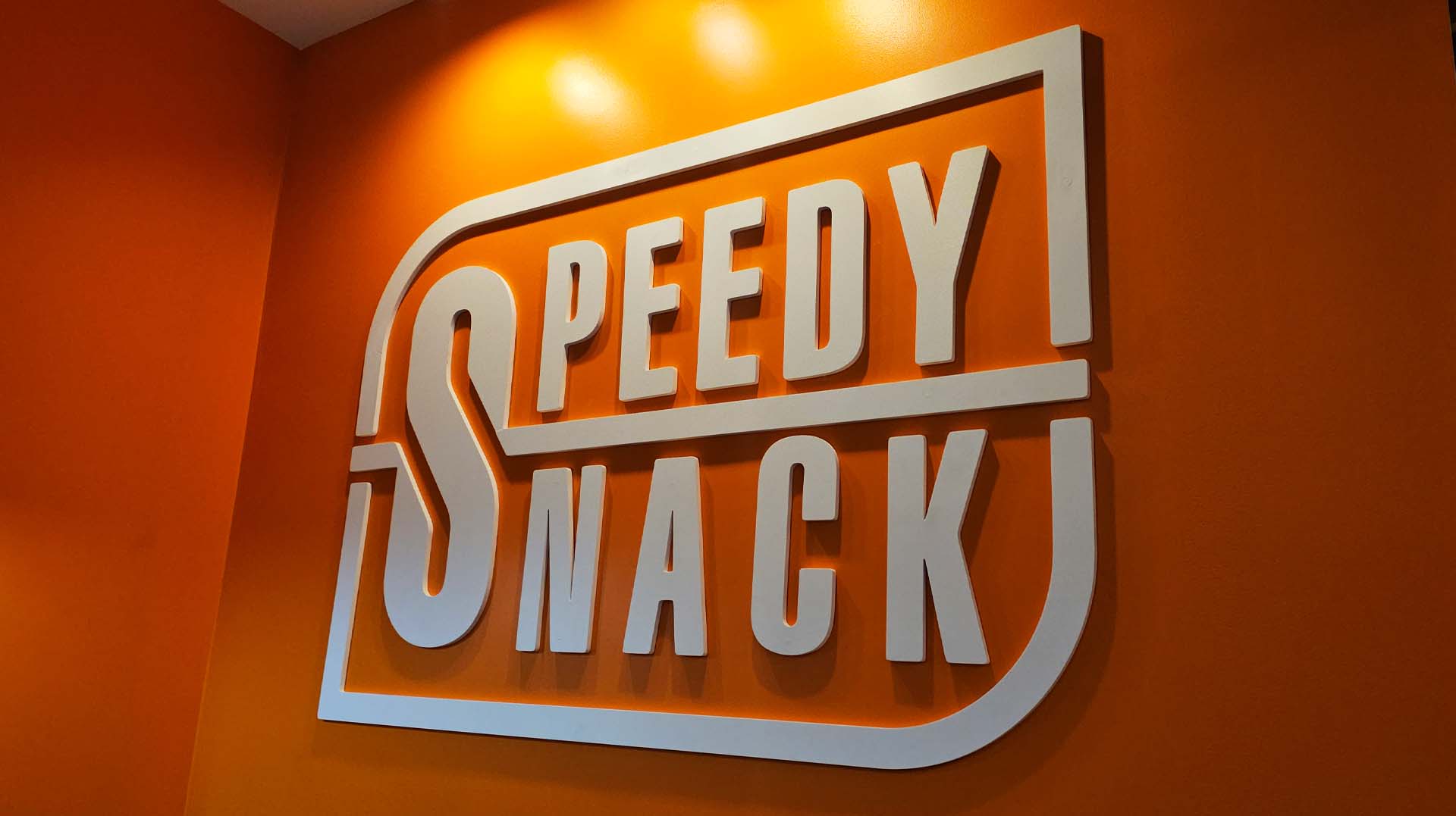 White 3D letters spelling 'SPEEDY SNACK' mounted on an orange wall.