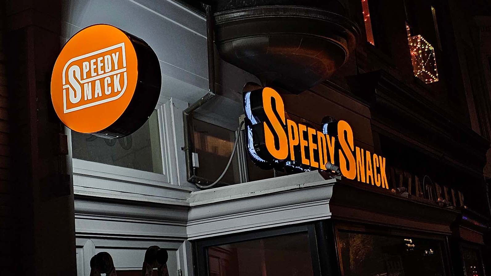 Illuminated orange signs displaying 'Speedy Snack' on the exterior of a building at night.