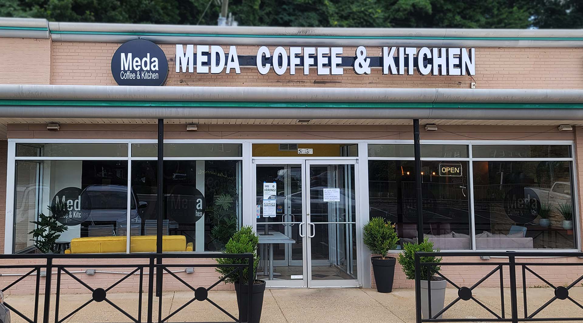 Front facade of Meda Coffee & Kitchen with glass doors, potted plants, yellow and beige couches visible inside, and an open sign lit.