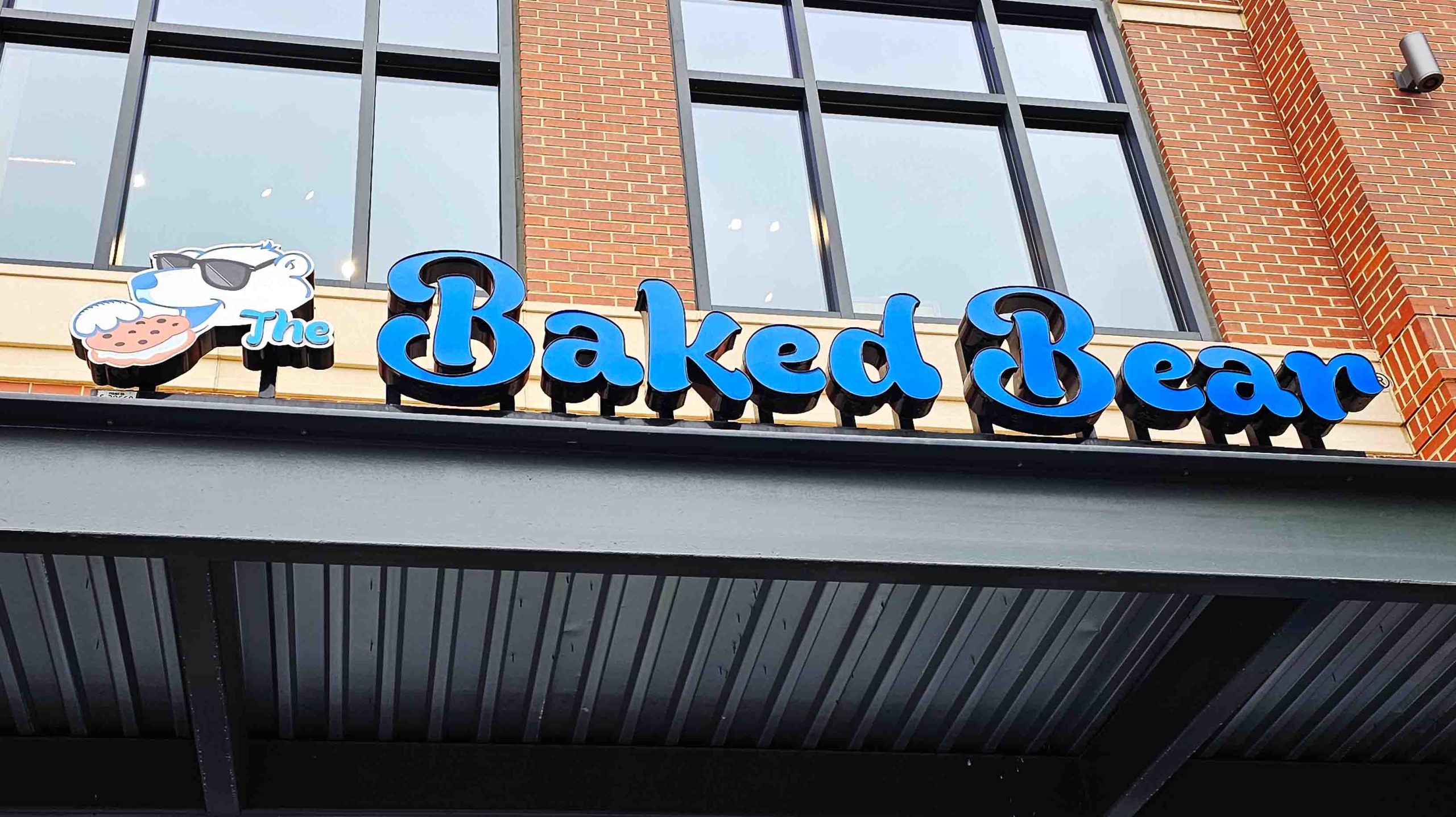 The Baked Bear store sign with a blue bear wearing sunglasses holding a cookie on a brick building.