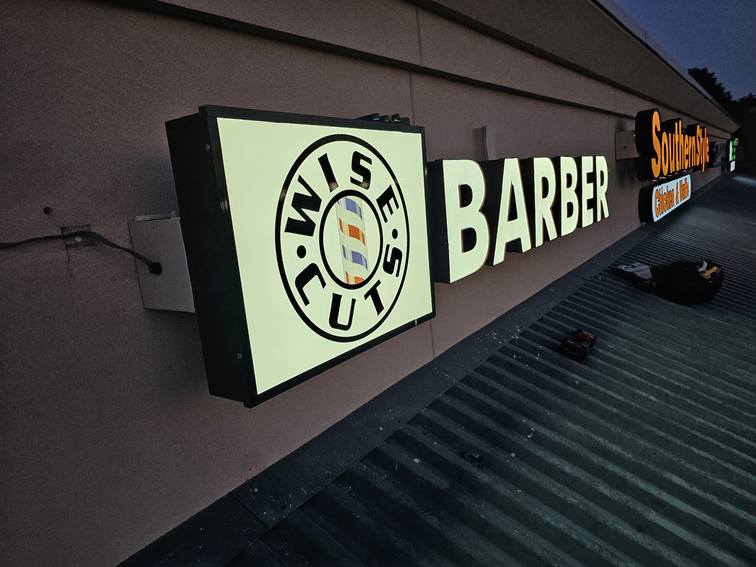 Illuminated barber sign with the words WISE CUTS and a barber pole icon on a building at dusk.