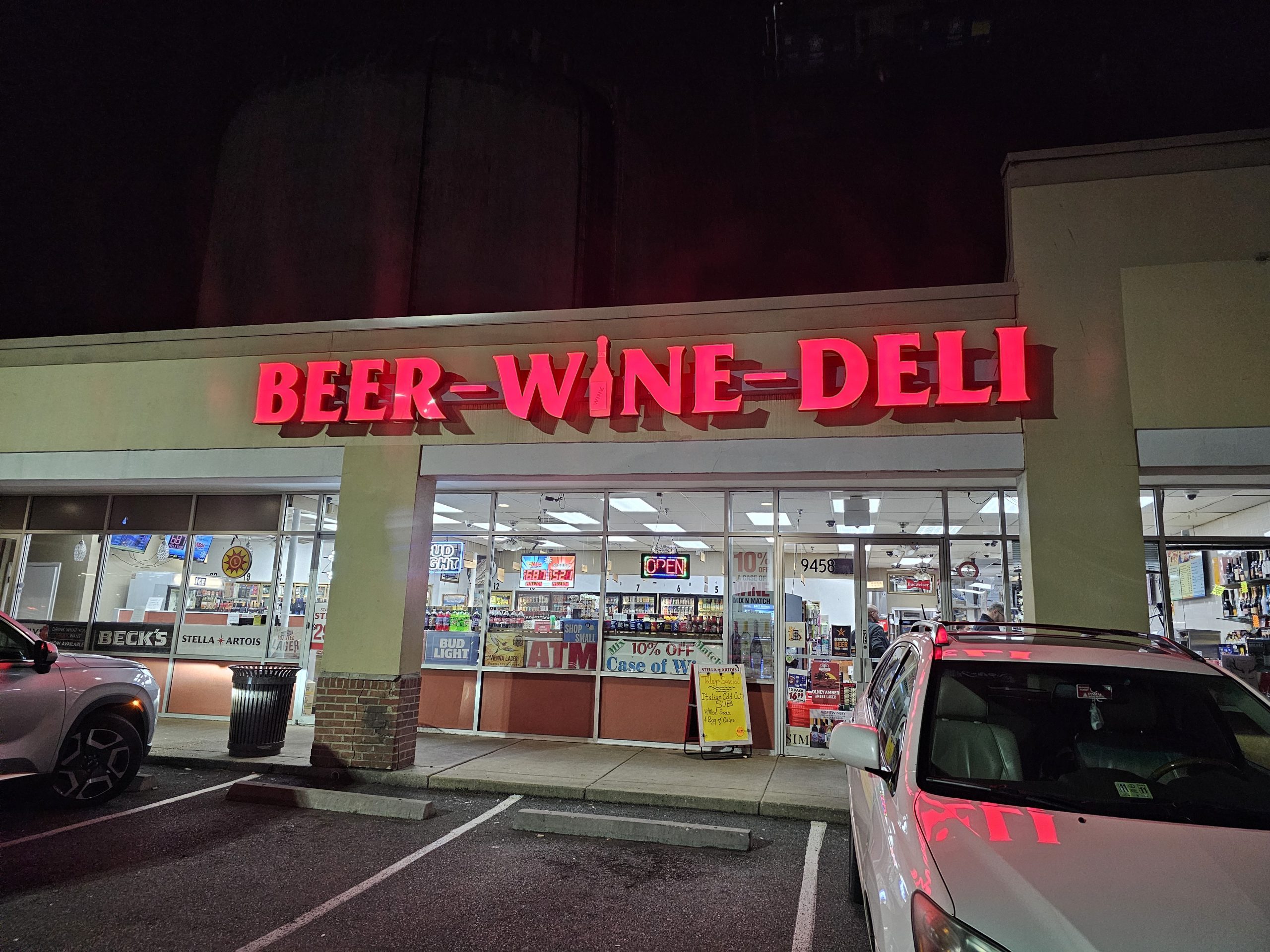 Store front at night with bright red neon sign reading 'BEER - WINE - DELI' and cars parked in front.