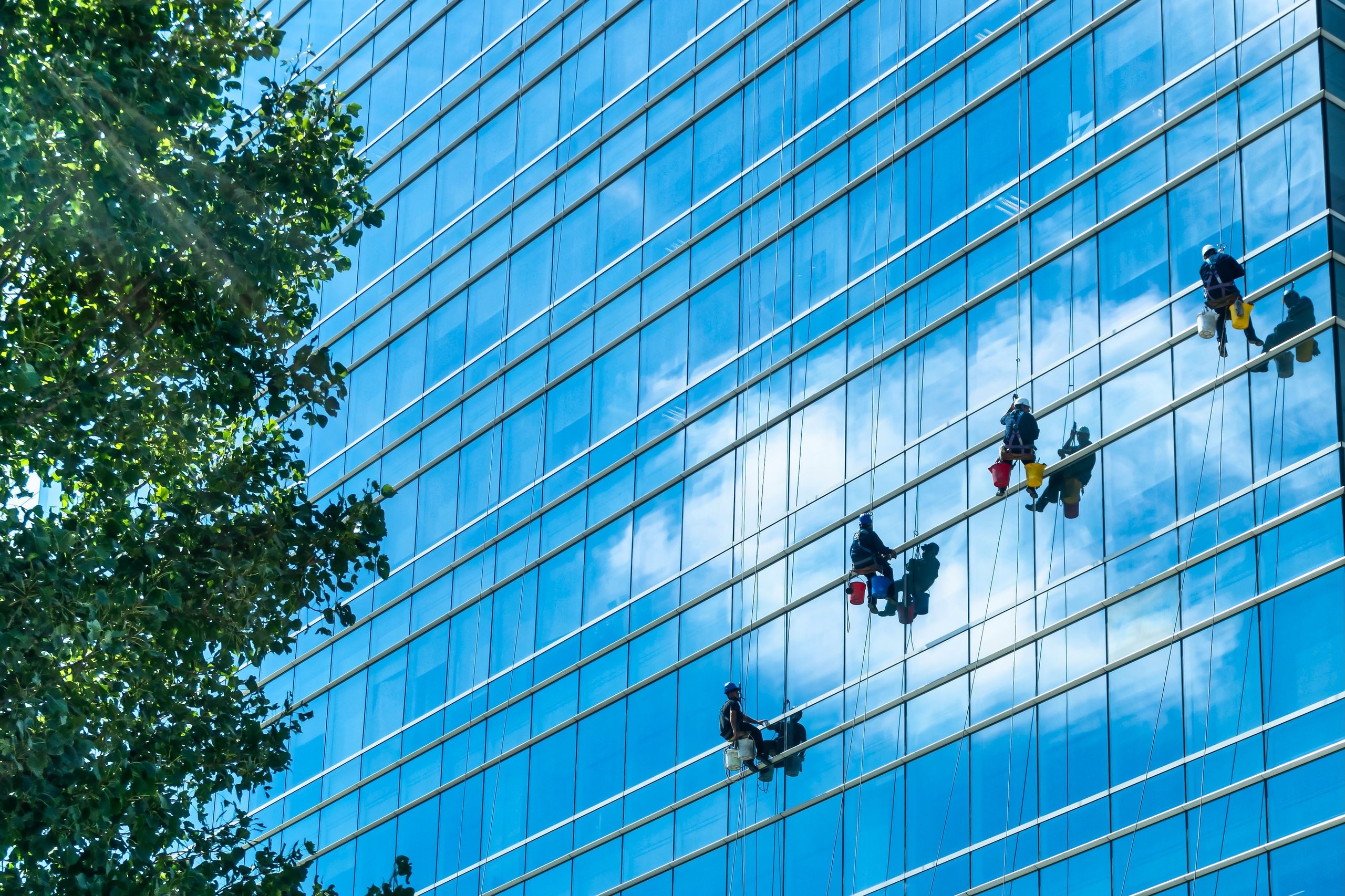 Three window cleaners suspended on ropes cleaning the glass facade of a modern skyscraper with reflections of clouds and blue sky.