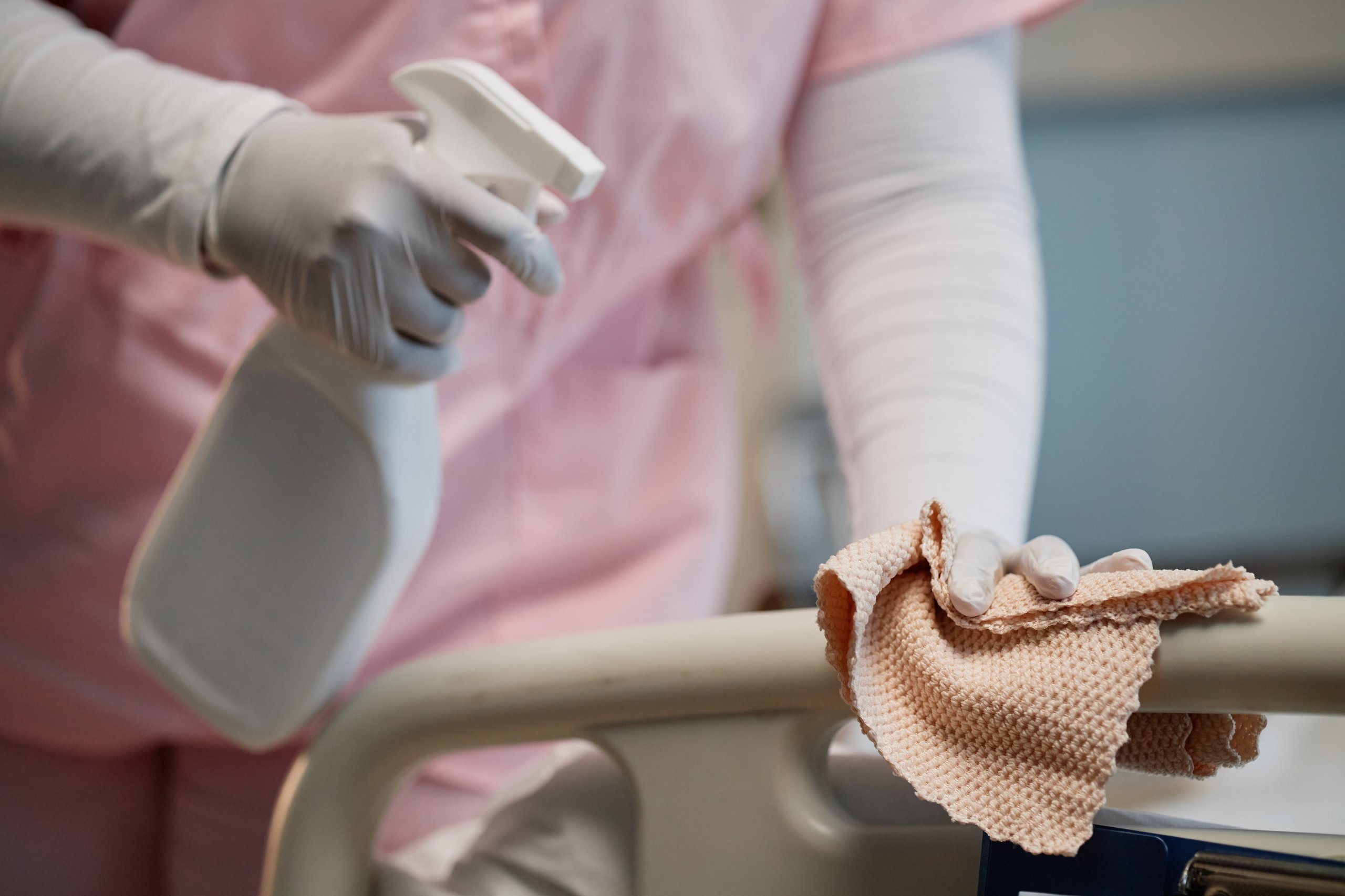 Person wearing gloves and pink scrubs cleaning a hospital bed rail with a spray bottle and cloth.