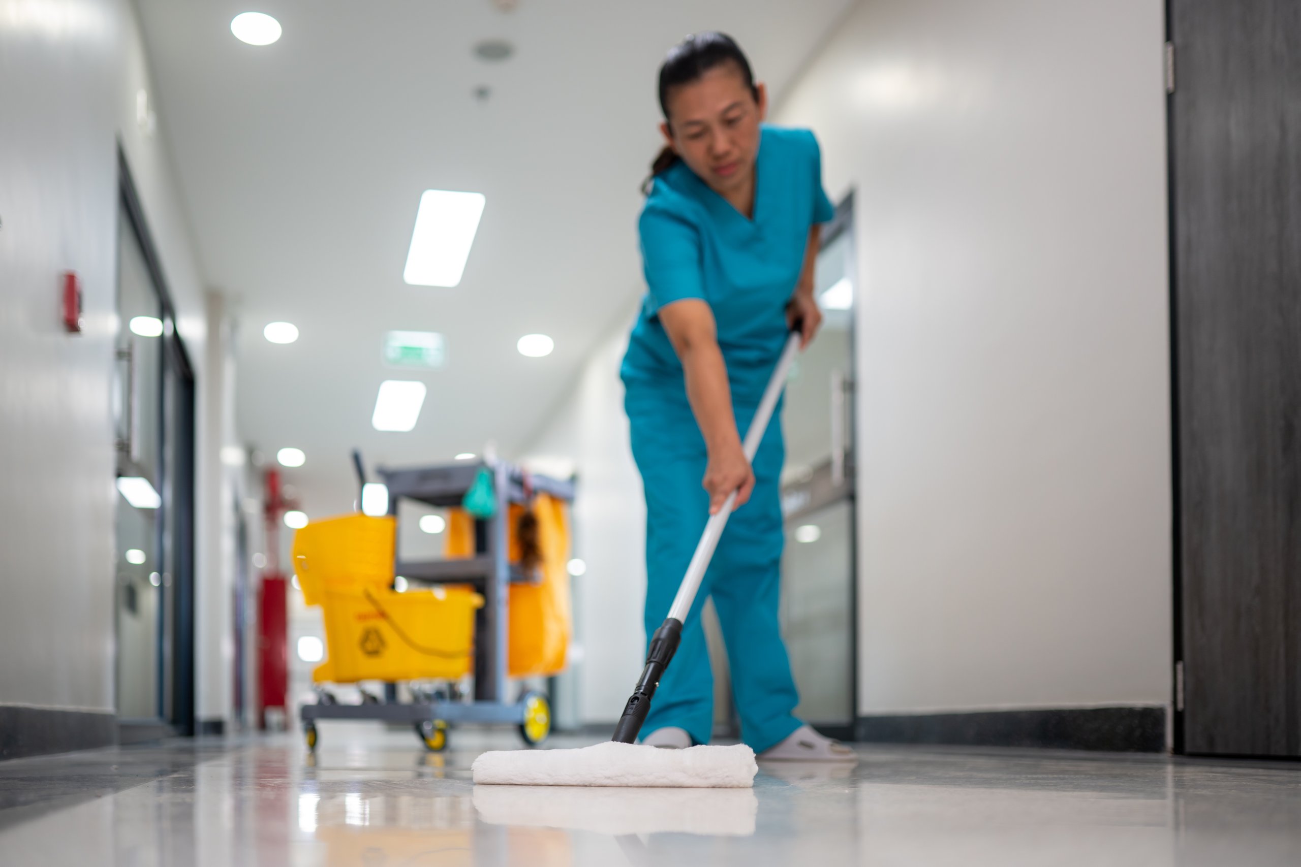 Janitor in blue scrubs mopping a shiny hospital hallway floor with a yellow cleaning cart in the background.