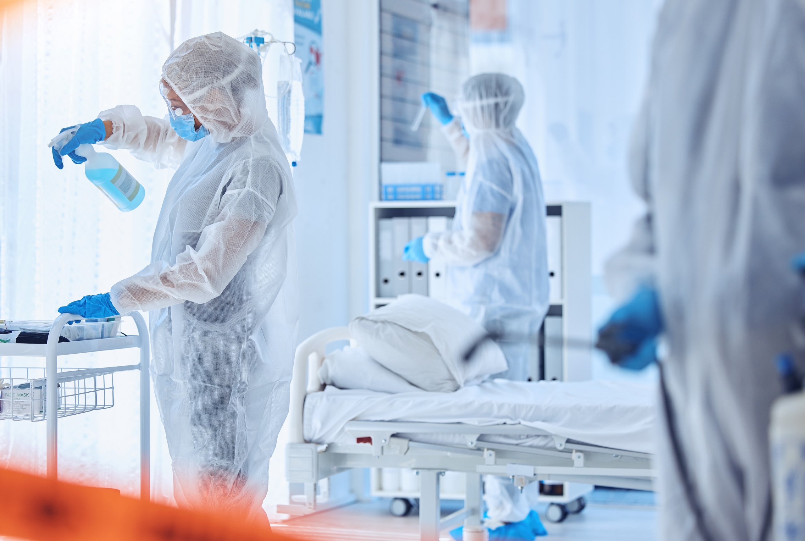 Healthcare workers in full protective suits sanitizing a hospital room with an empty patient bed.
