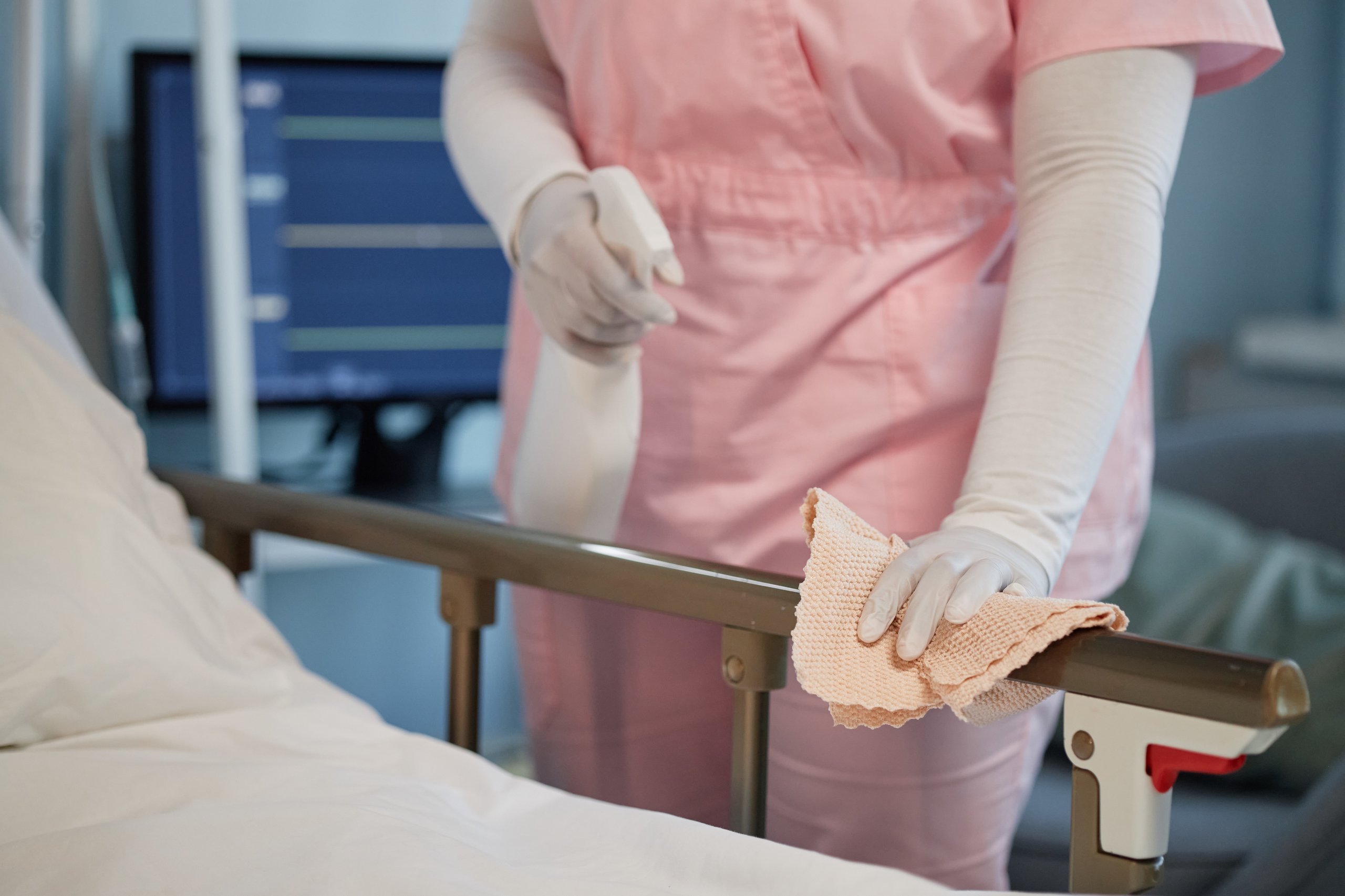 Healthcare worker wearing gloves and pink scrubs disinfecting a hospital bed rail with a spray bottle and cloth.