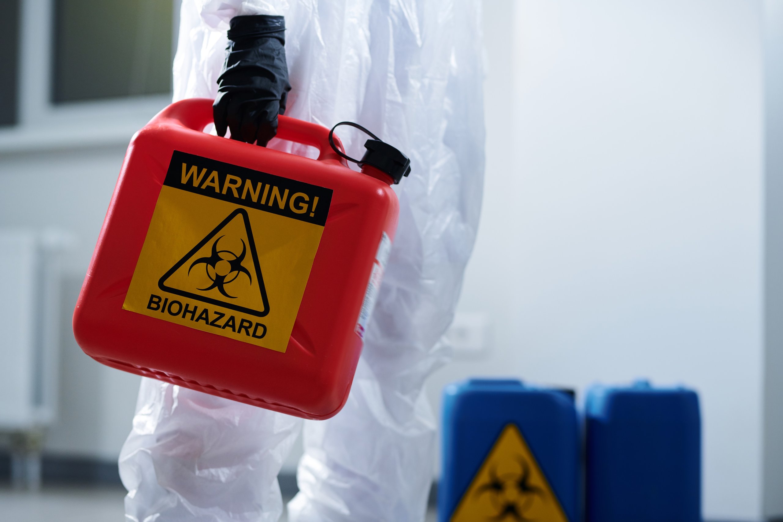 Person in protective suit holding a red container labeled with a yellow biohazard warning sign.