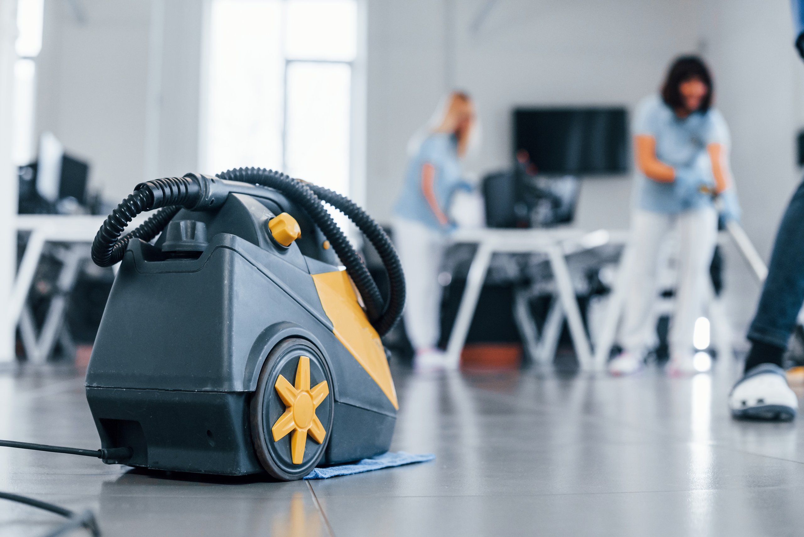 Close-up of a floor cleaning machine with yellow wheels on a tiled floor in an office setting with blurred people cleaning in the background.