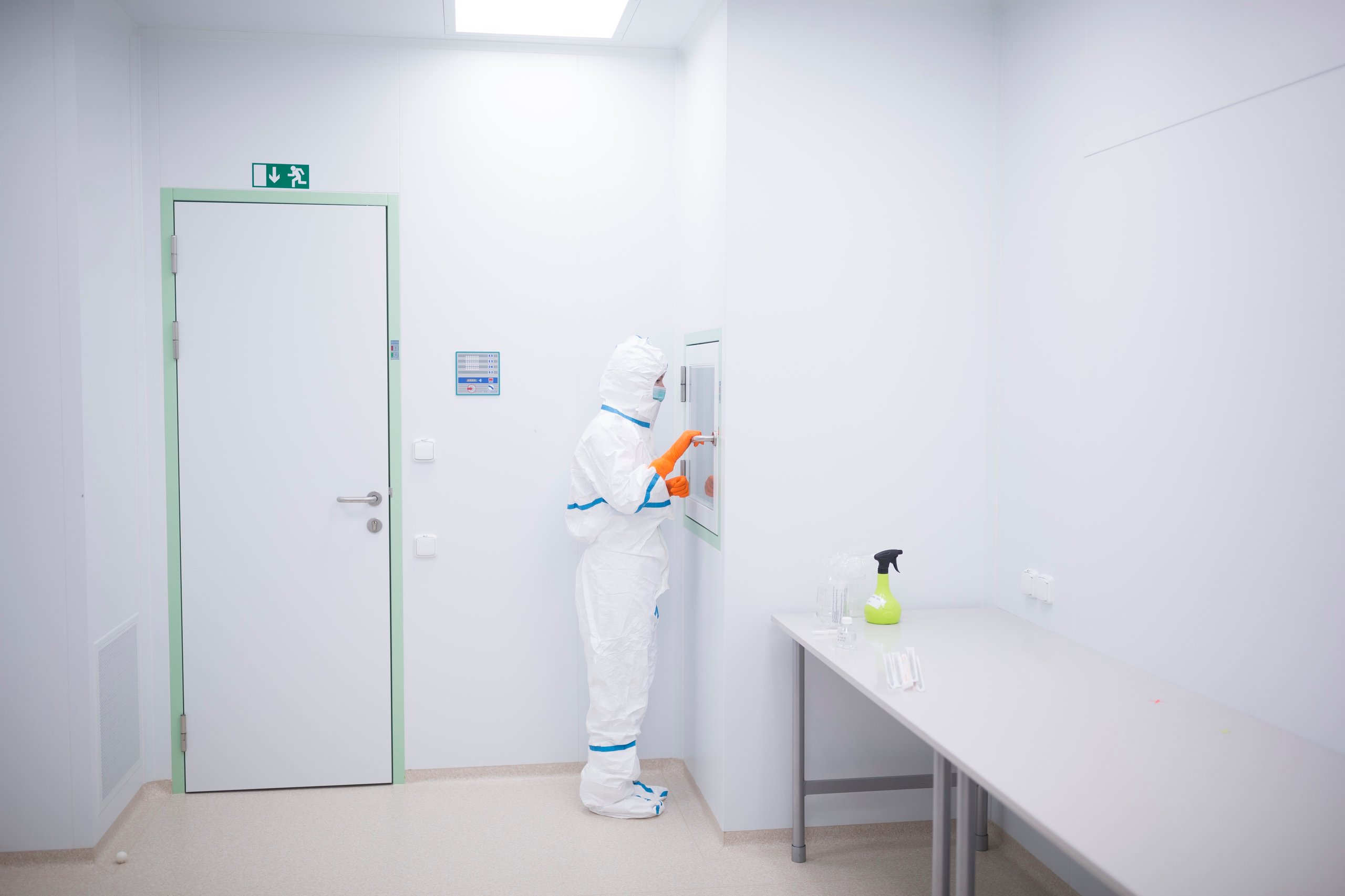 Person in full protective suit with orange gloves operating a control panel inside a clean, sterile room.