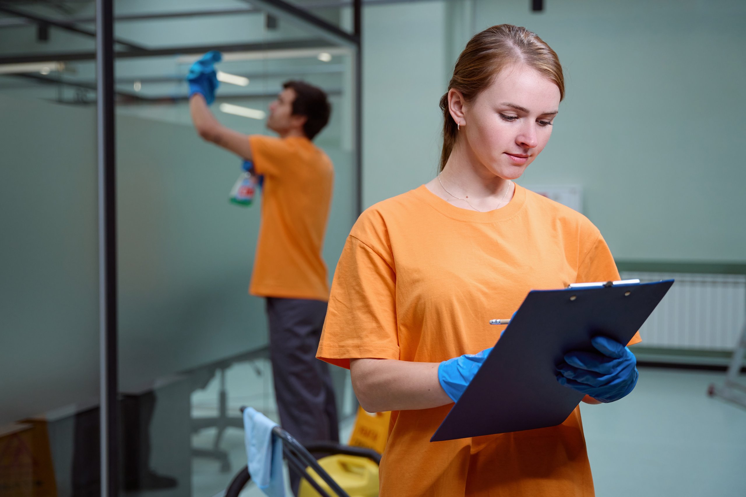 Woman in orange shirt and blue gloves holding a clipboard while a man cleans a glass wall in the background.