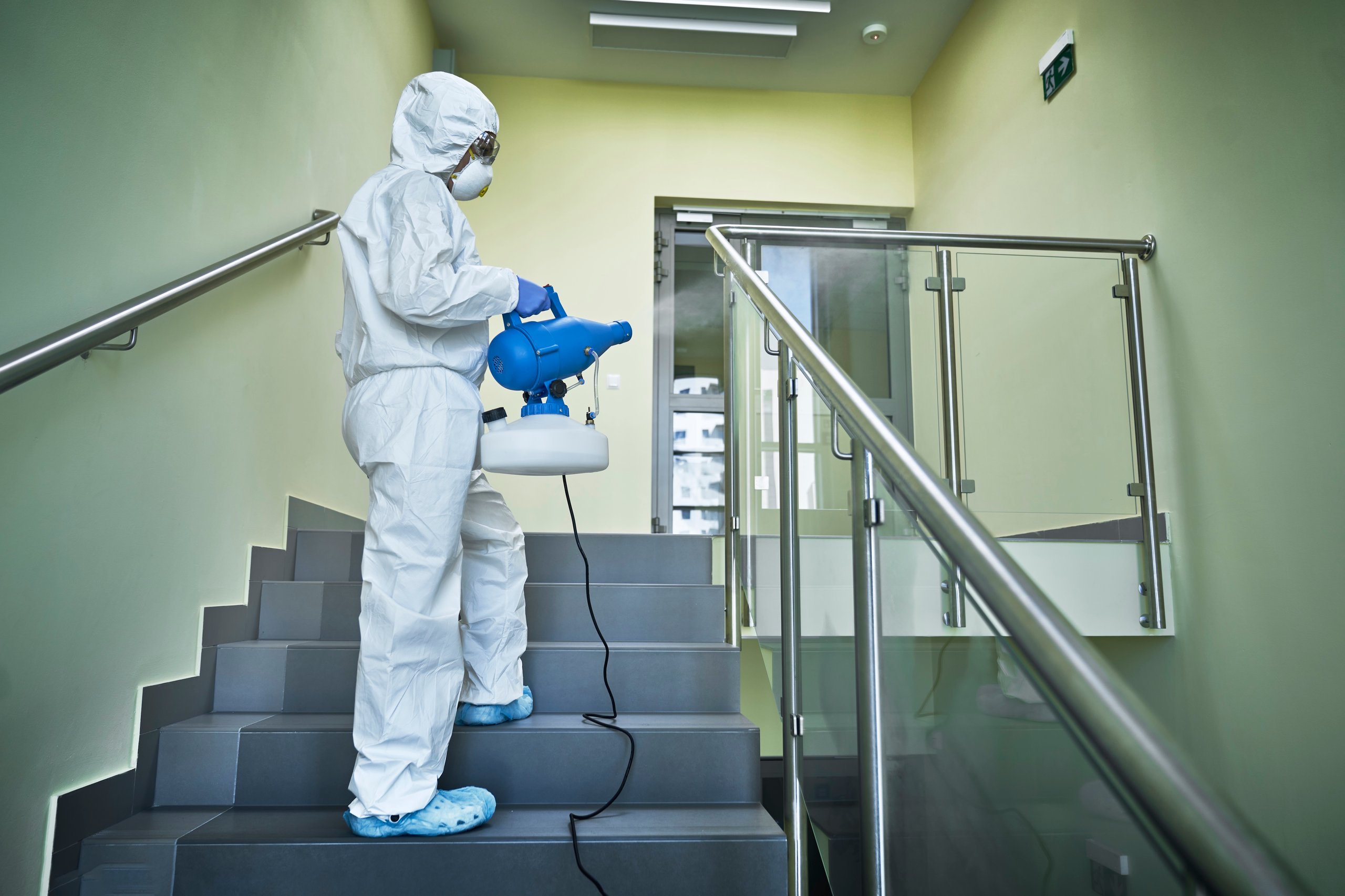 Person in full protective suit disinfecting stairway with a blue fogging machine.