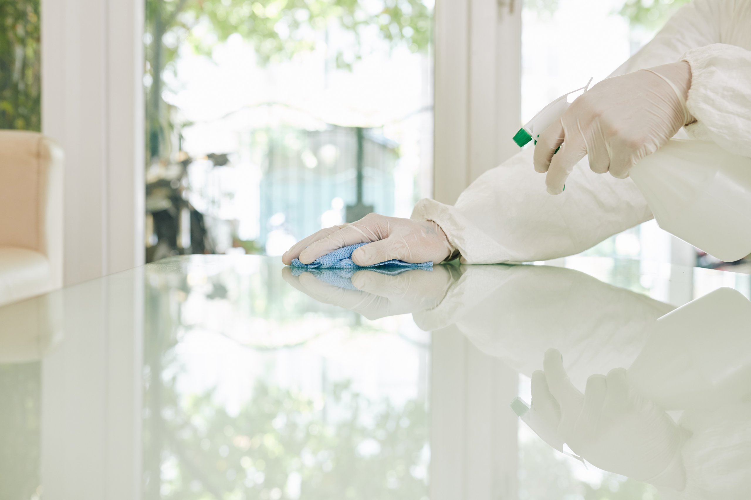 Person wearing white protective clothing and gloves cleaning a glass table with a blue cloth and spray bottle.