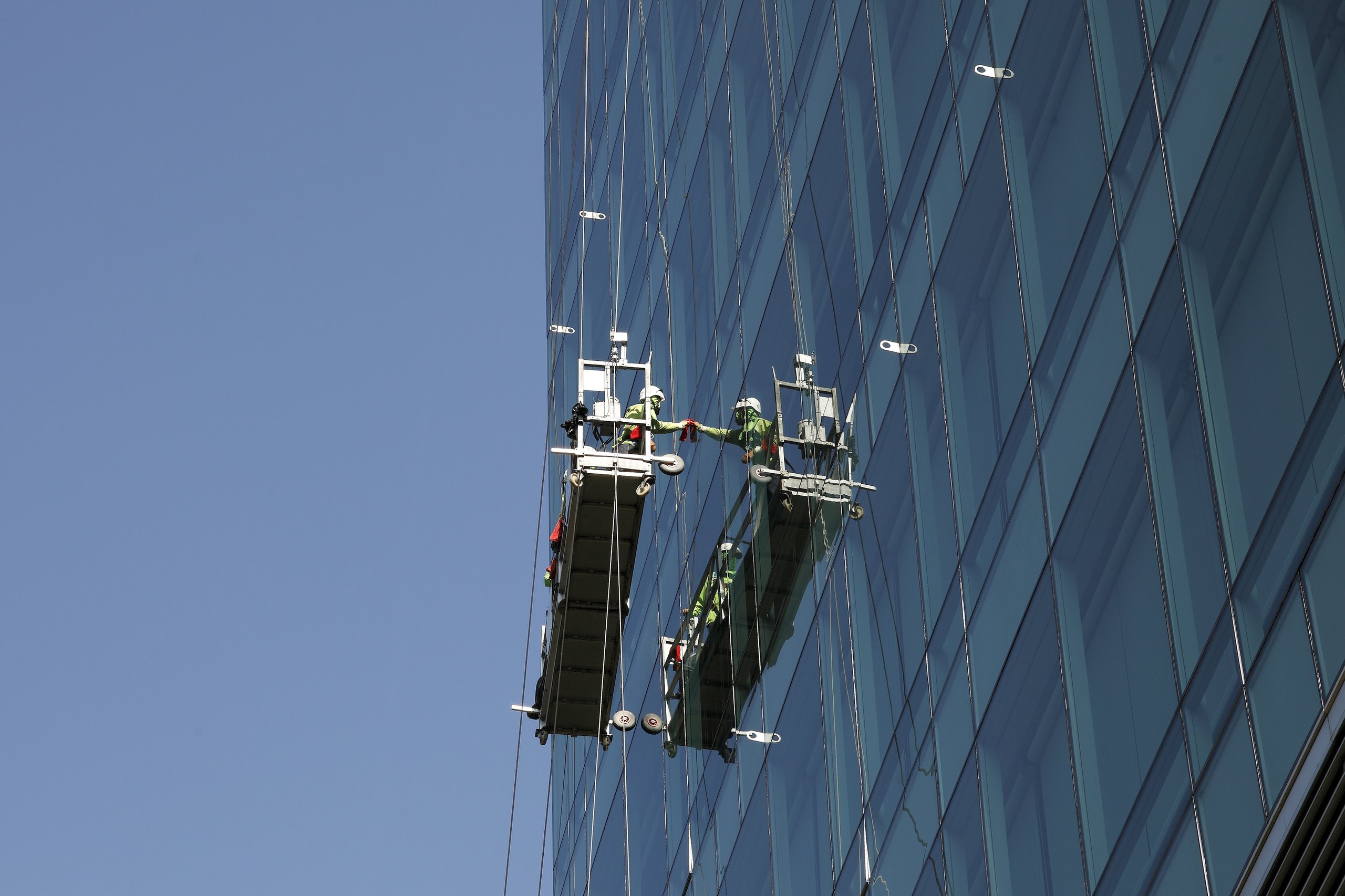Two window washers on suspended platforms cleaning a glass skyscraper against a clear blue sky.