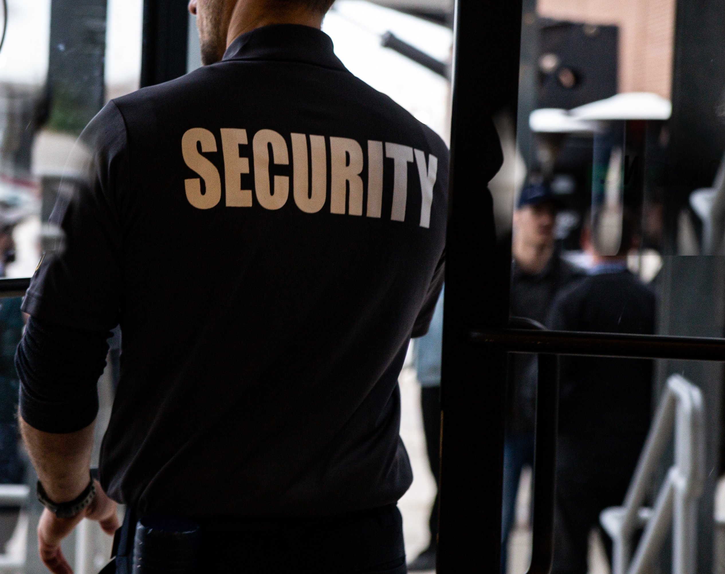 Rear view of a security guard wearing a black uniform with SECURITY written in bold white letters, standing near a glass door.